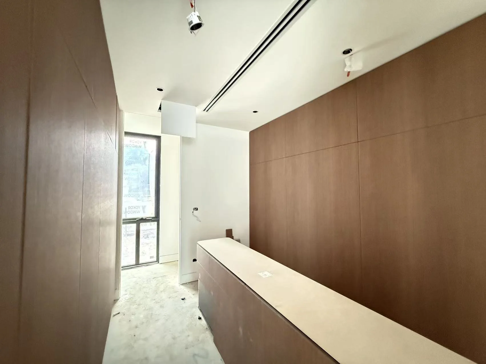 Interior of a room under construction with wooden-paneled walls, a window, and a ceiling with recessed lighting and hanging electrical wires.