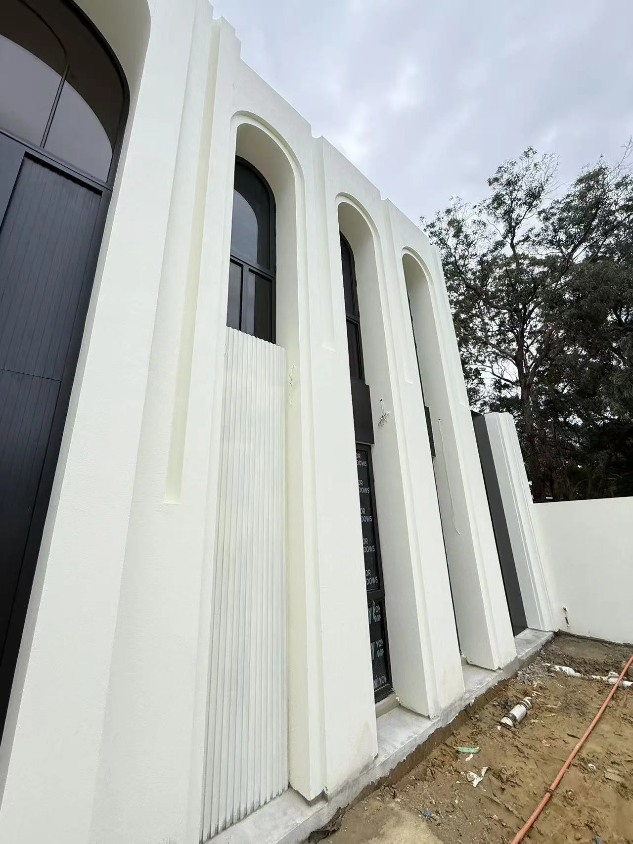 Modern white building with tall arched windows and decorative vertical panels, under construction with exposed ground and construction pipes.