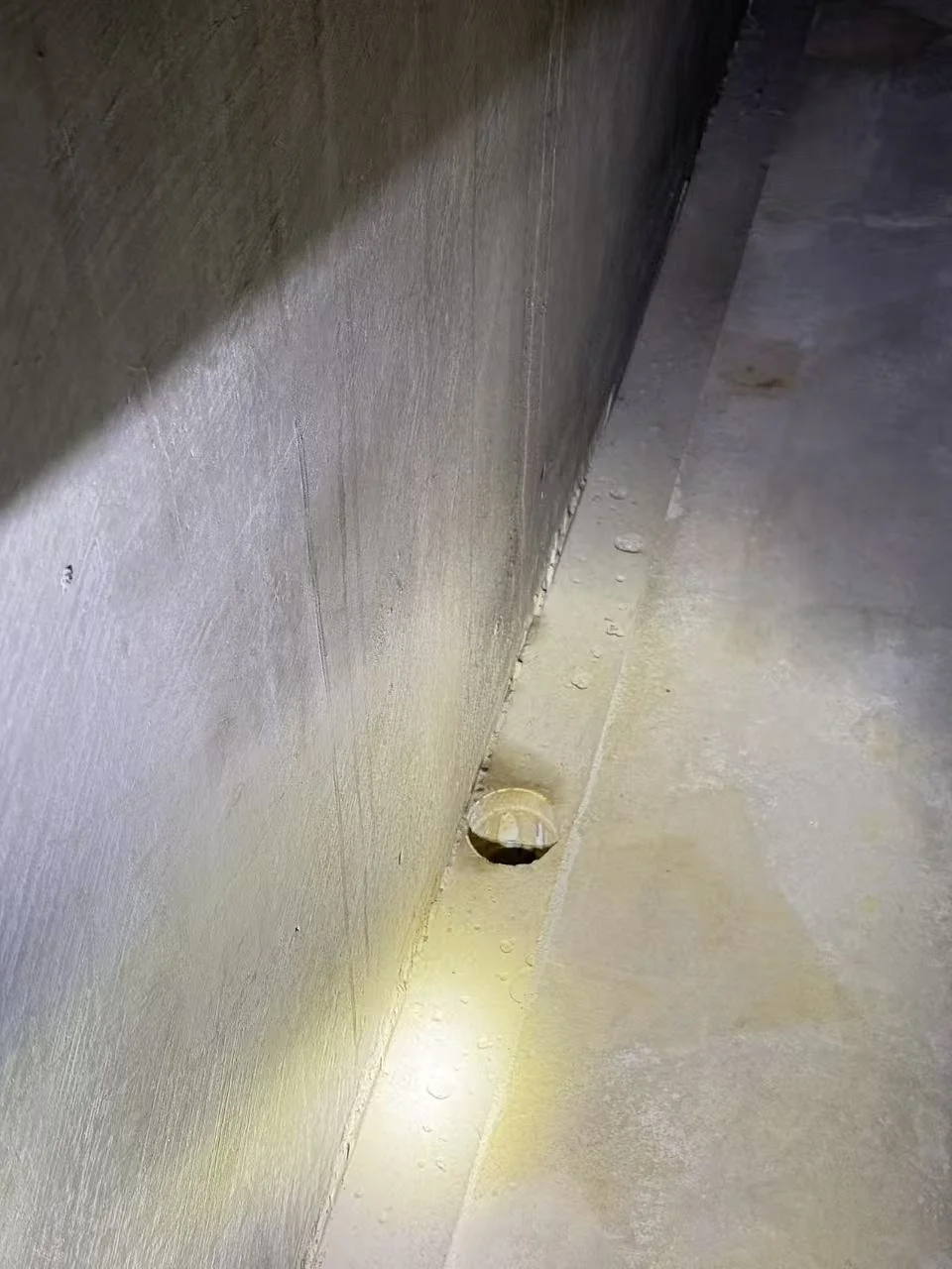 Close-up of a corner on a floor with a small drain or vent on the floor near the base of a wall.