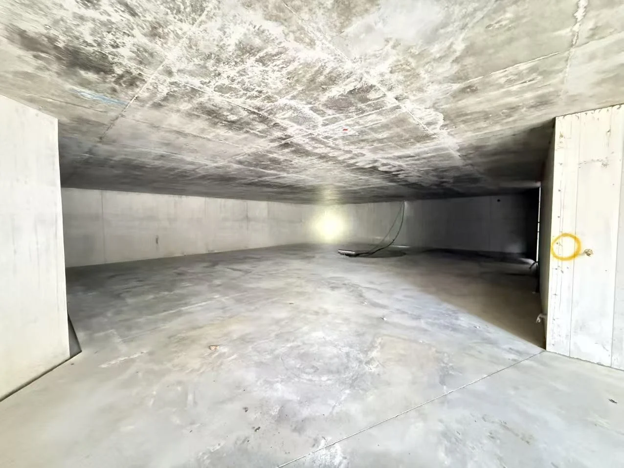 Underground parking garage with concrete walls, floor, and ceiling, showing cables hanging from the ceiling and a yellow lock on one wall.