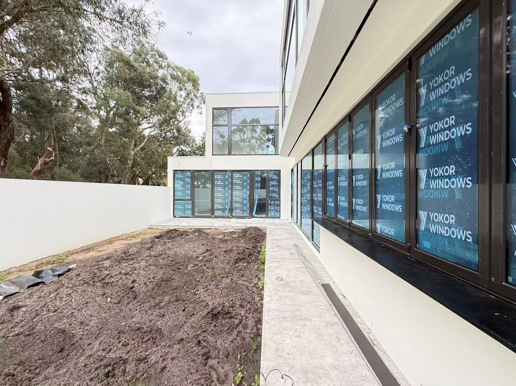 New building with large windows covered with blue protective film displaying 'Yokor Windows' in white, some trees, and an overcast sky.