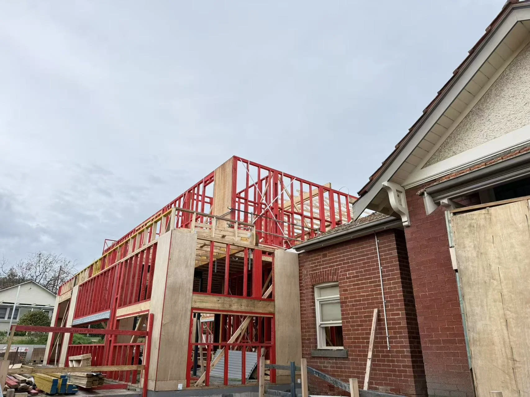 Construction of a new extension on a brick house with red framing and unfinished wooden sections under a cloudy sky.