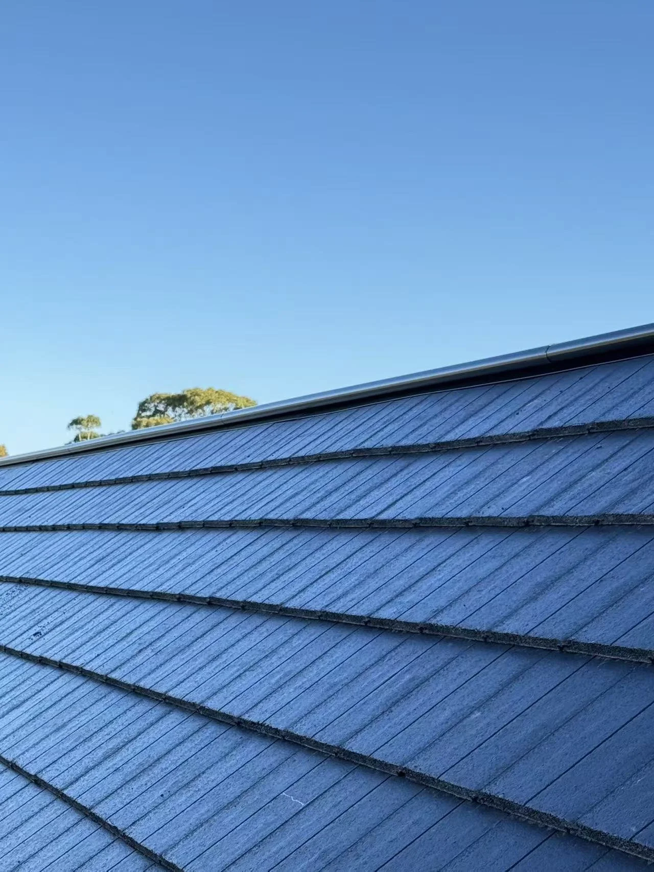A close-up view of a metal roof with overlapping panels, set against a clear blue sky with some trees in the background.