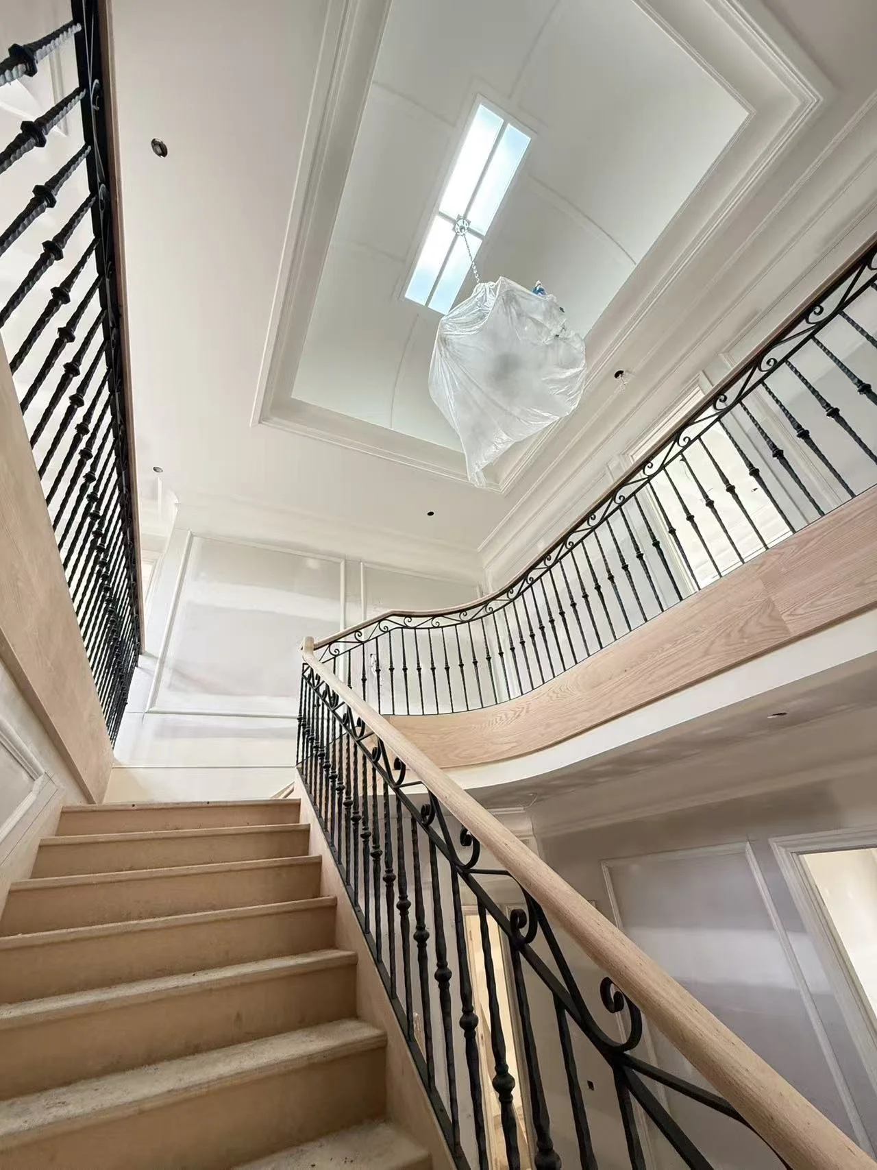 The image shows a staircase inside a house with light wood steps and black iron railings, leading to a mezzanine. The ceiling features a large decorative tray design with a skylight and a hanging plastic bag, possibly containing construction or renov