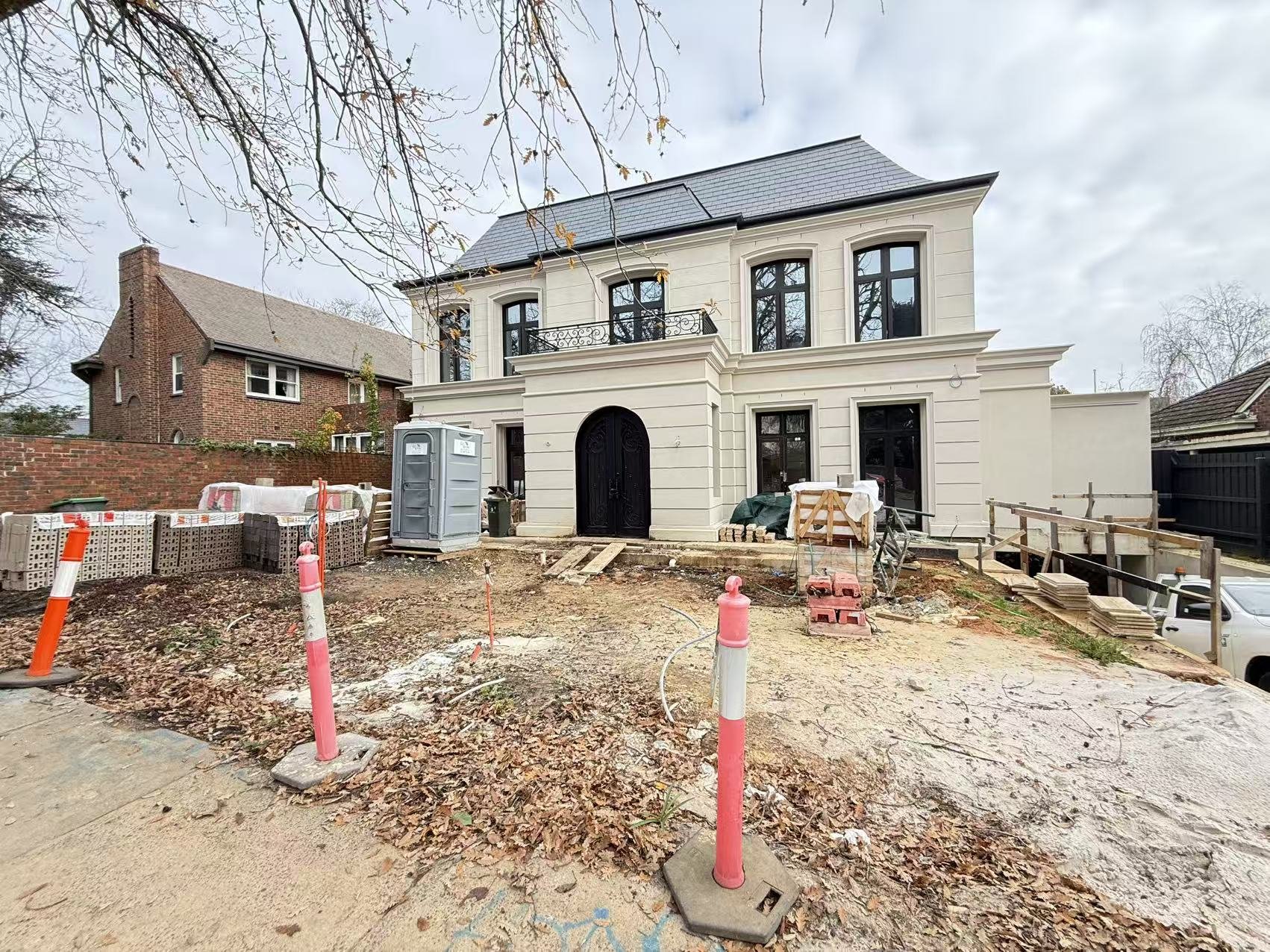 Under construction house with a black front door, white exterior walls, and black window frames, construction materials and tools in front, surrounded by orange cones and protective barriers.