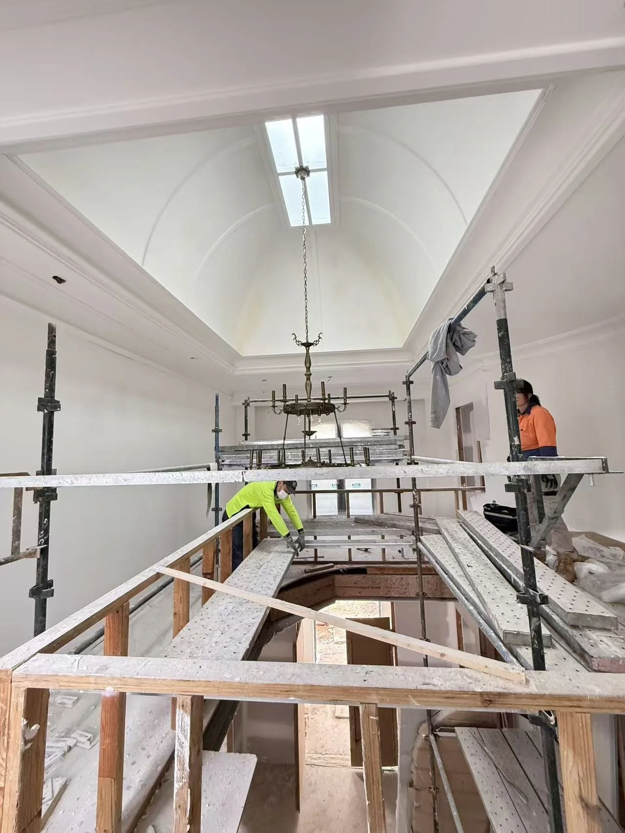 Construction workers installing flooring on a scaffold inside a building with a high ceiling and chandelier.
