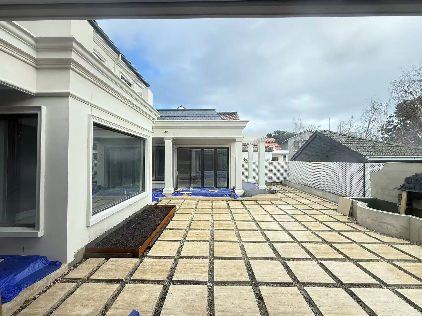 View of a backyard patio under construction with stone slabs arranged in a grid pattern. The house has white walls, large windows, and a sliding glass door. Strings of construction materials and tools are visible on the ground, with a partly cloudy s