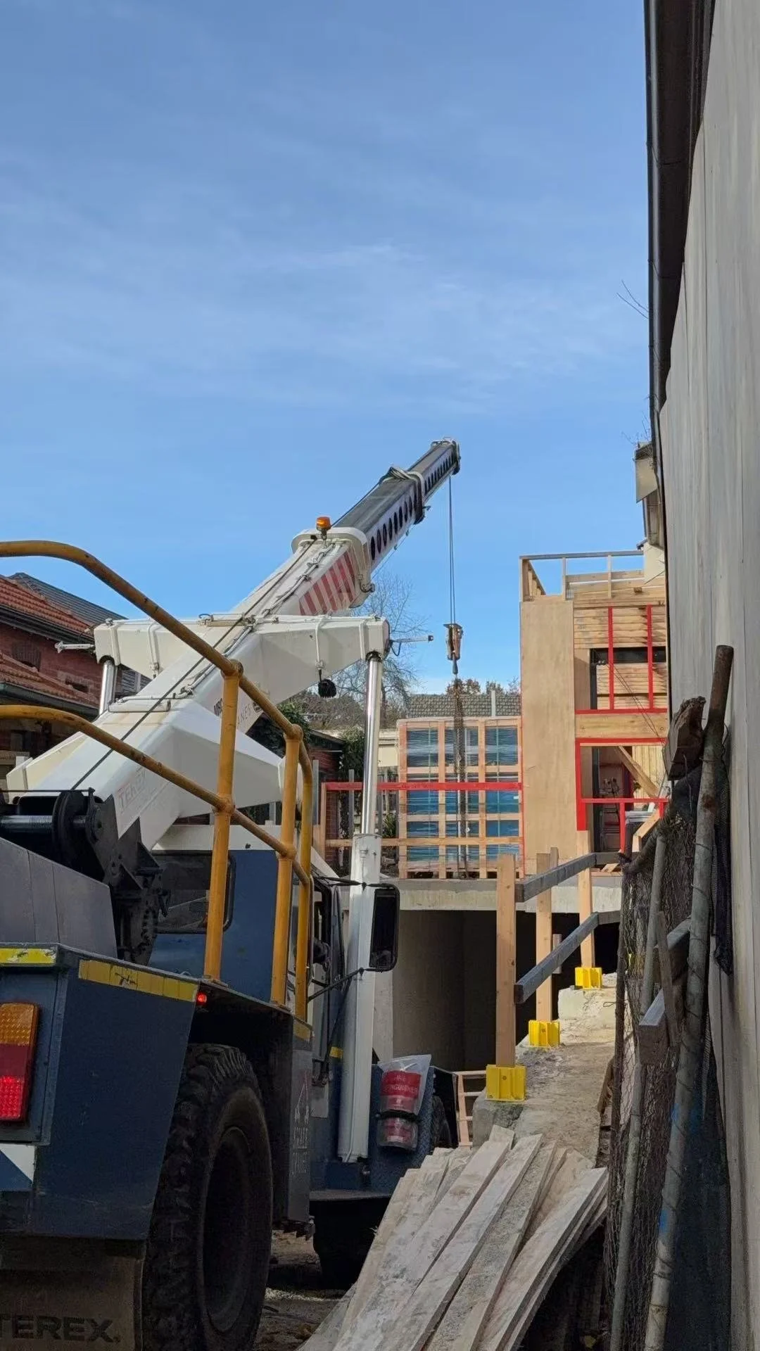 Construction site with a crane lifting materials, partially built wooden structures, and building materials on the ground.