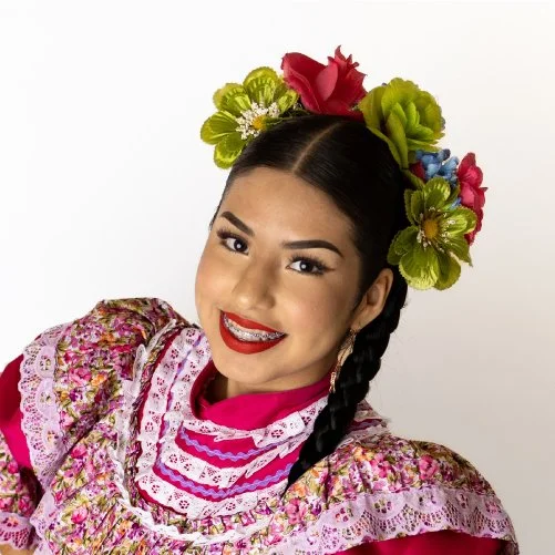 Young woman dressed in traditional Mexican folkloric attire with a floral headpiece and braid, smiling at the camera.