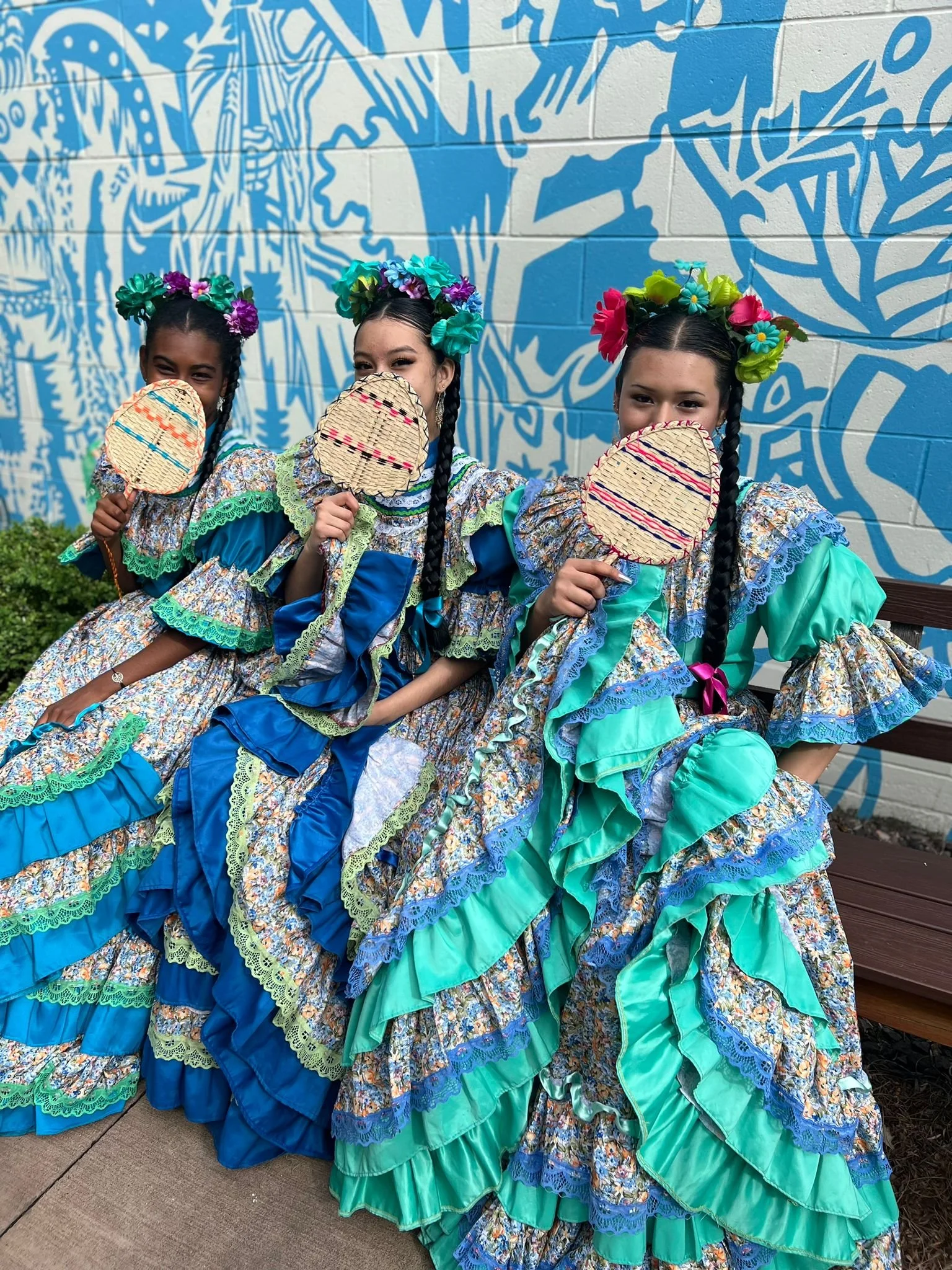 Three girls in colorful traditional Mexican dresses with floral headpieces, holding woven fans, sitting outdoors against a blue mural background.
