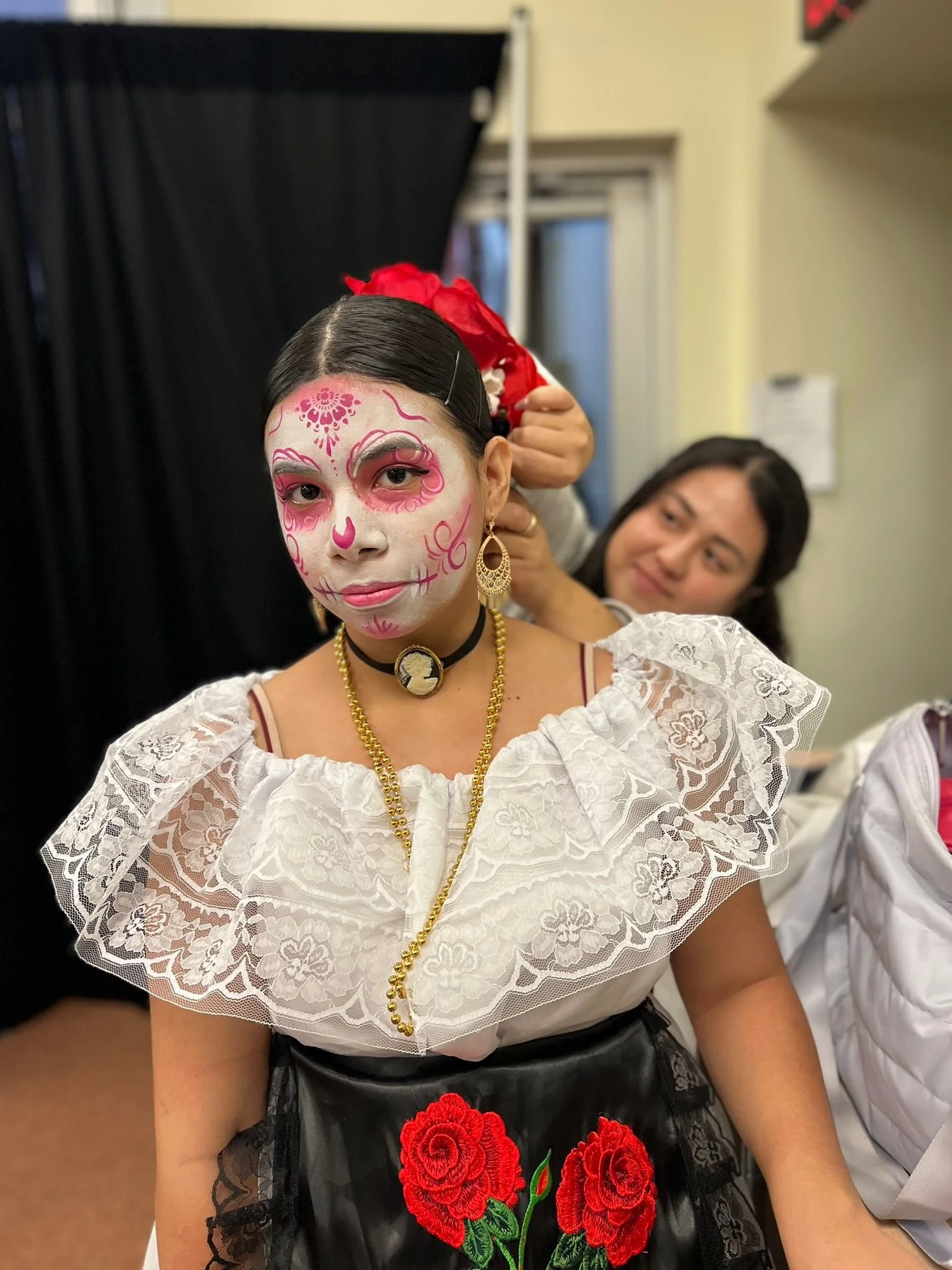 Young woman with sugar skull makeup, traditional dress, and jewelry, getting her hair styled by a woman in the background.