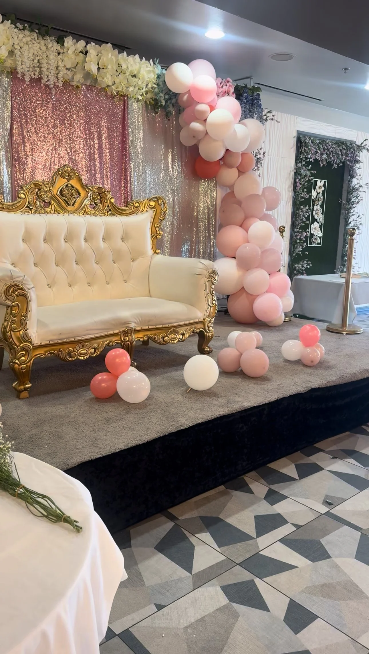 Elegant decorated stage with cream and gold vintage sofa, pink and white balloons, floral backdrop, and tables for a celebration or event.