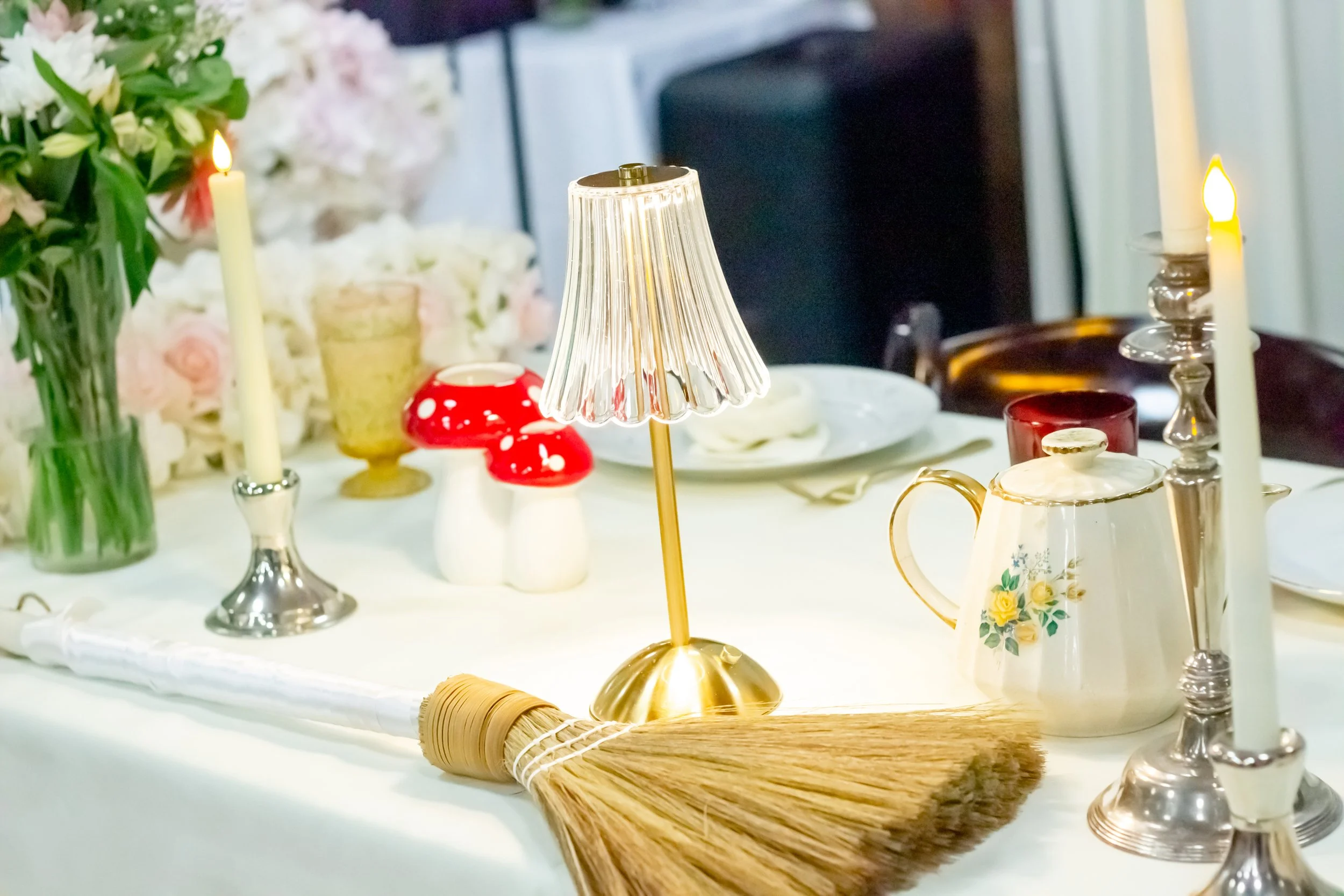 Close-up of a dining table set with candles, a floral centerpiece, a tea pot, a small lamp, red mushroom-shaped decor, and a broom, with plates and glasses in the background.