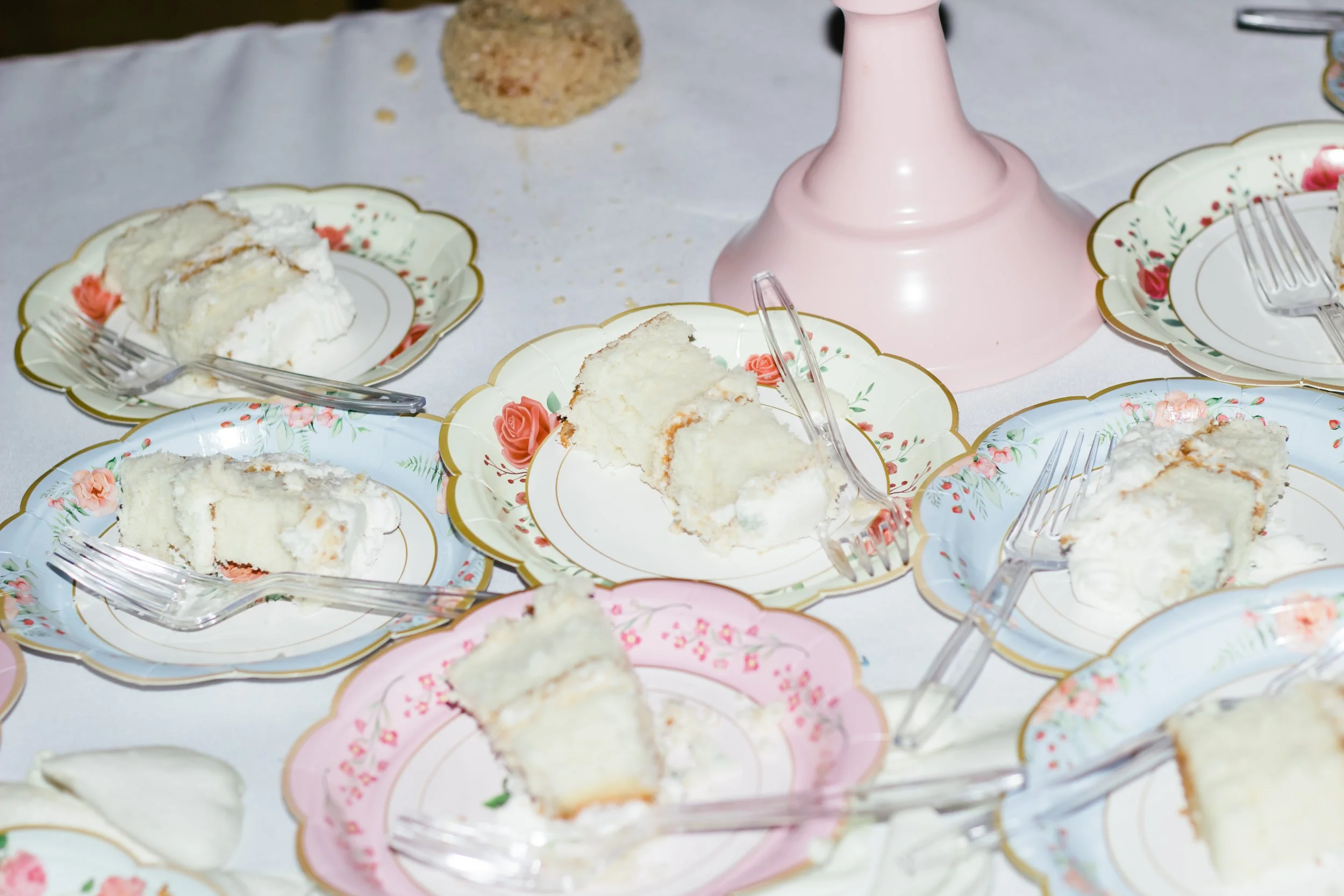 Pieces of white frosted cake on vintage floral plates with forks, set on a white tablecloth with a pink cake stand in the background.