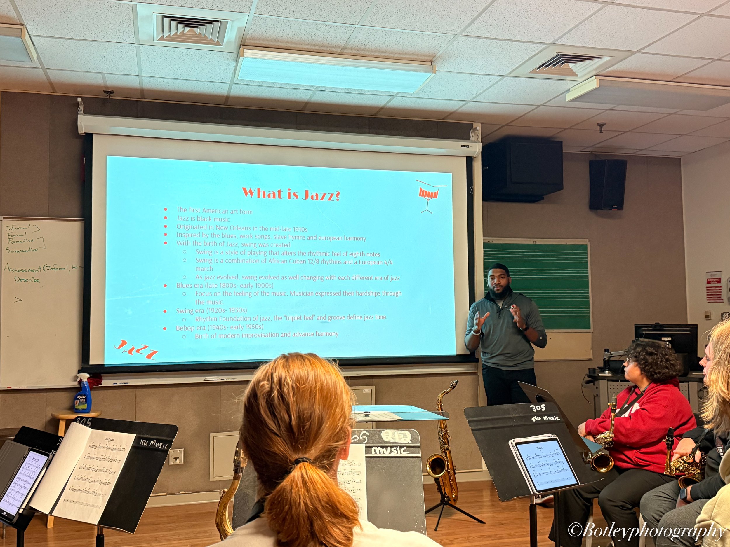 A classroom setting with a music instructor giving a lecture on jazz, featuring a projector screen with a presentation slide titled 'What is Jazz?' and students sitting with musical instruments such as saxophones and music stands.
