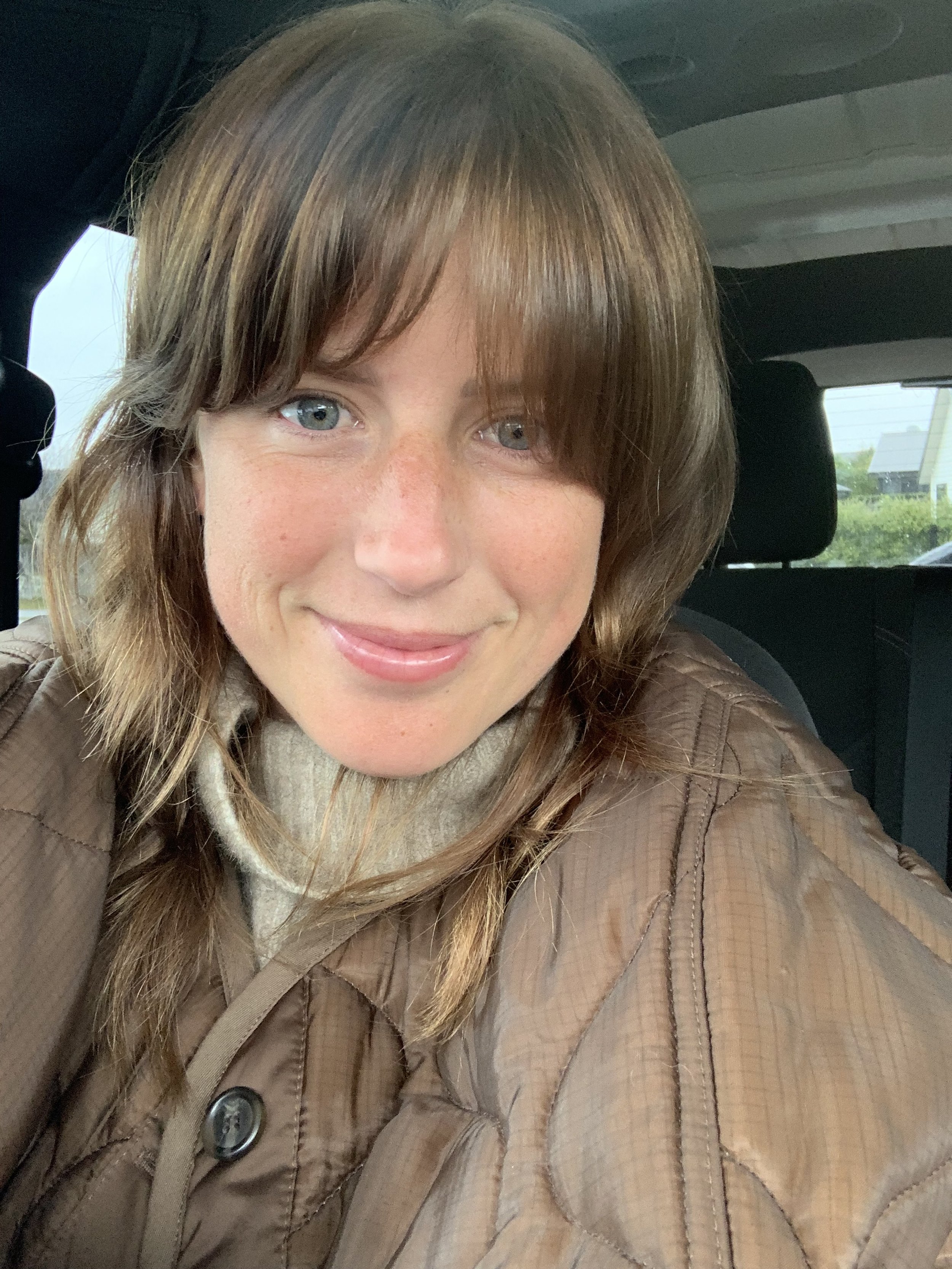 Close-up of a smiling woman with brown hair, blue eyes, and freckles, wearing a brown jacket inside a vehicle with a suburban neighborhood visible outside the window.