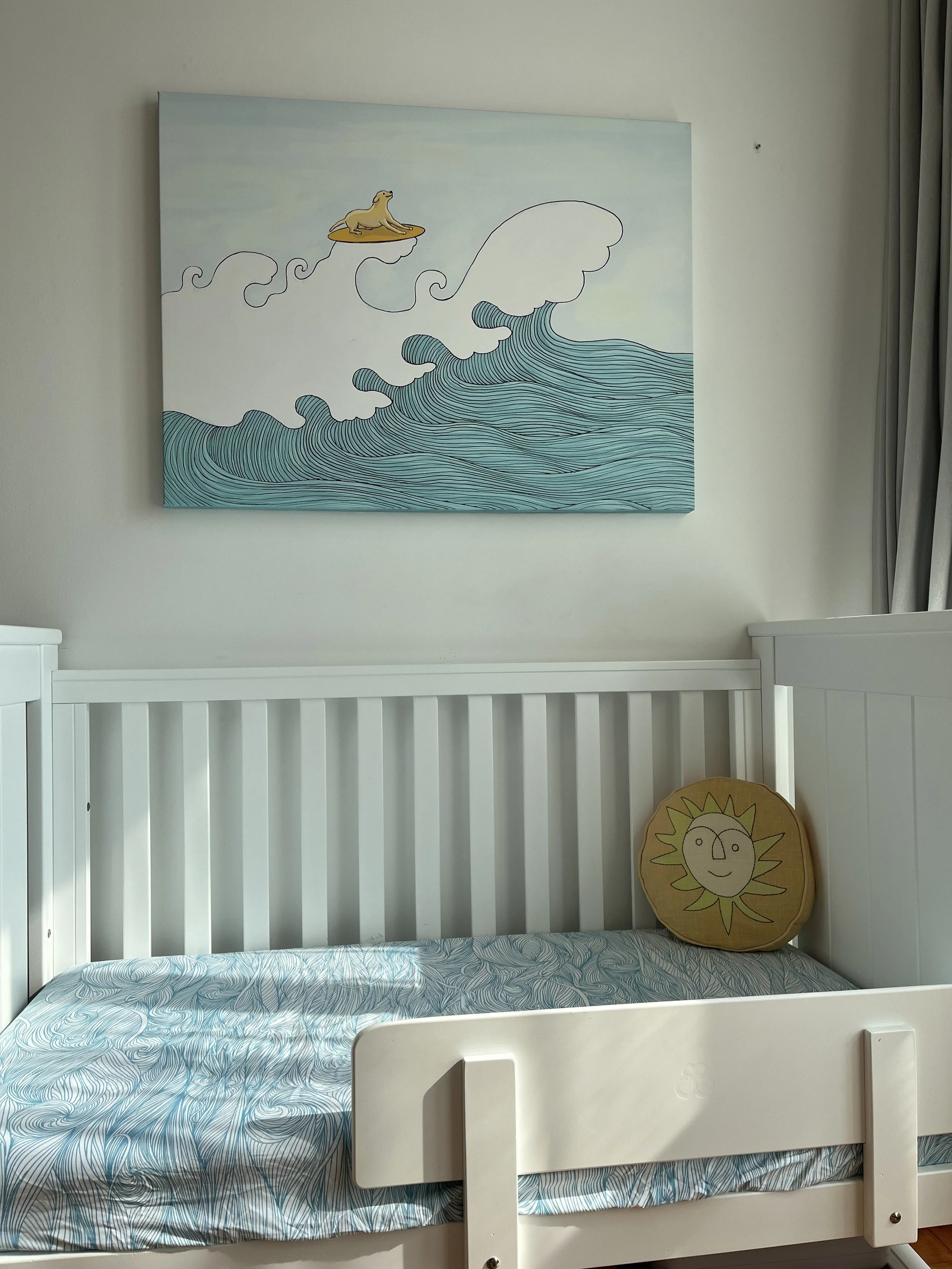 A white crib with a blue swirl patterned bed sheet and a round pillow with a sun face design, located in a room with a large ocean-themed wall art above it featuring a sea lion on a surfboard and a wave.