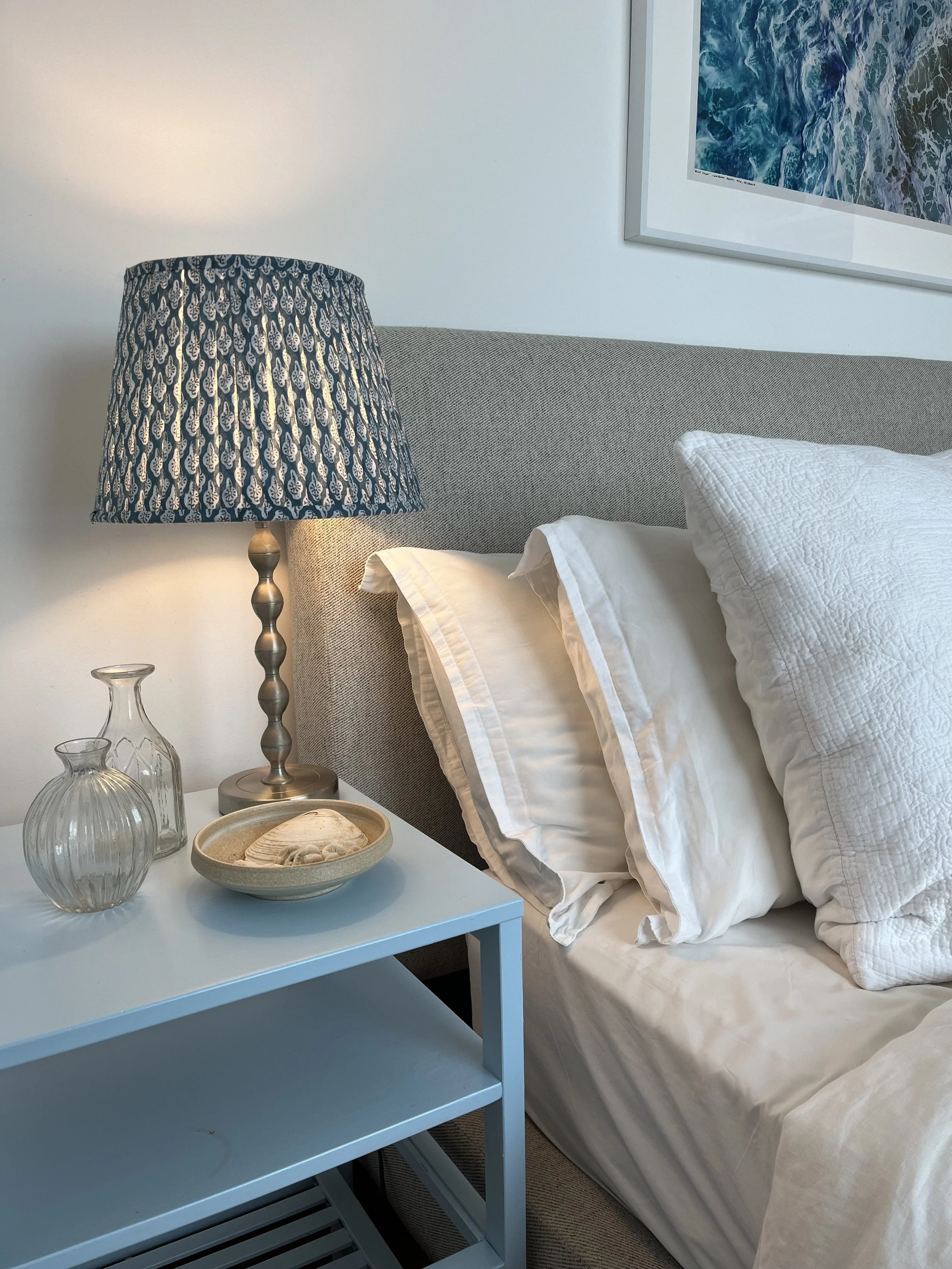 A bedside table with a glass vase, a smaller glass, and a bowl with shells. A lamp with a patterned shade is on the table next to a bed with white pillows and a gray headboard. Part of a framed picture of ocean waves is on the wall.