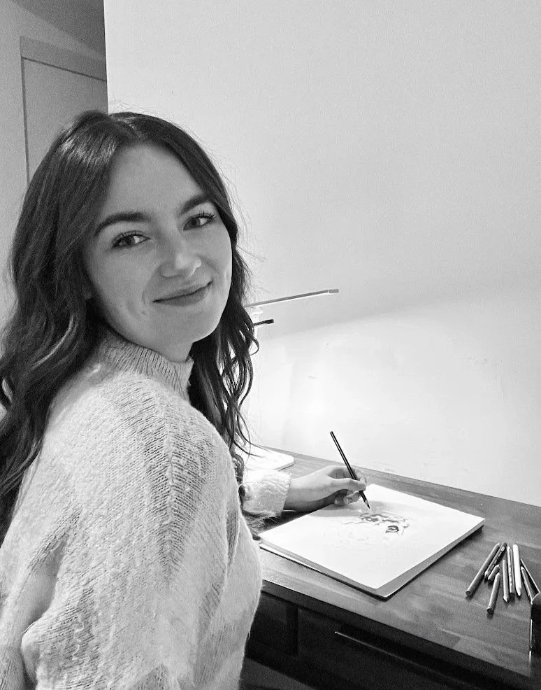 Summer Martens, artist, smiling while drawing on a dog at her desk in her room.
