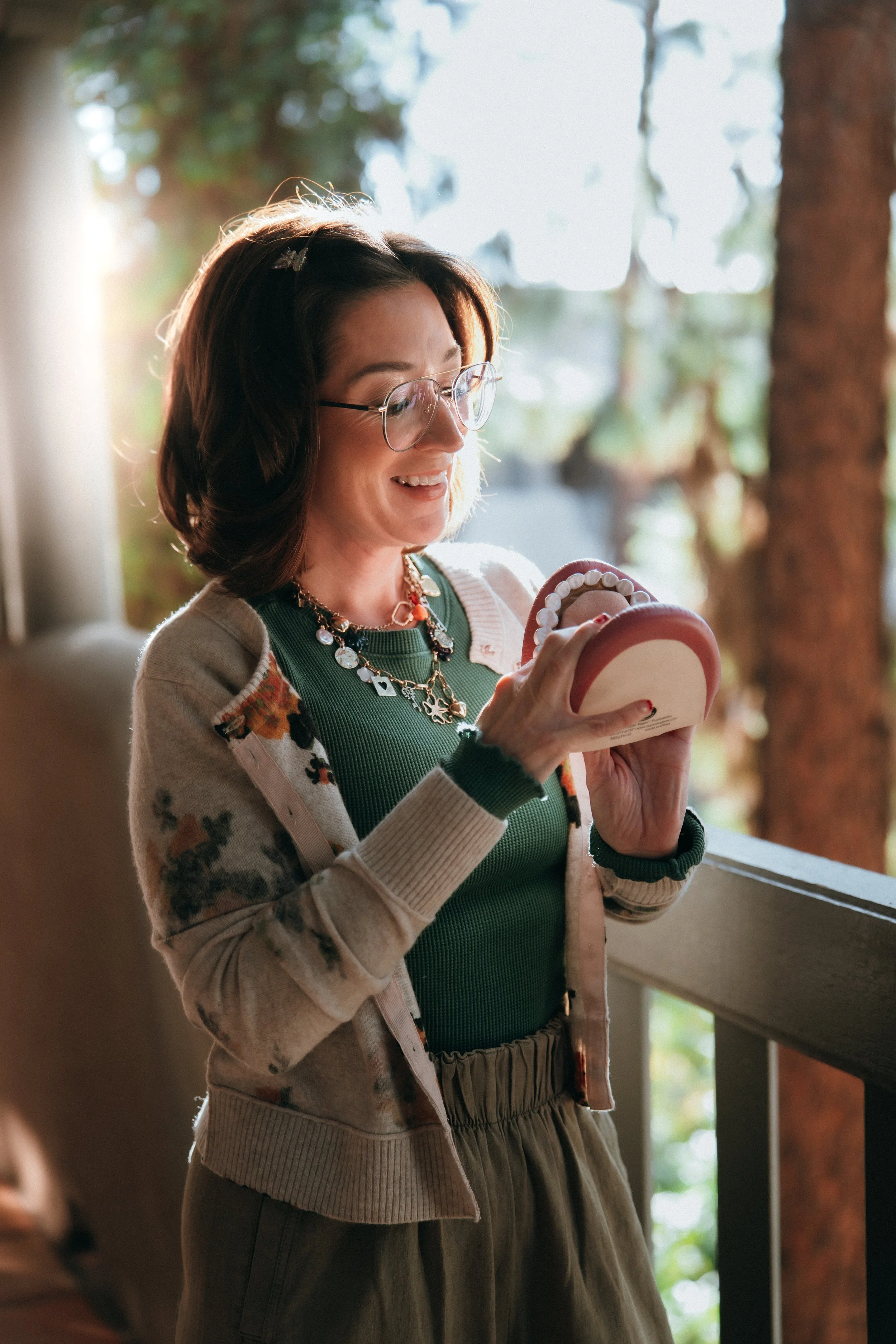 A woman with shoulder-length dark hair, wearing glass glasses, a green top, and a floral cardigan, smiling while looking at a mirror she holds in her hands. She is standing near a window with sunlight streaming in, with a blurred outdoor background visible.