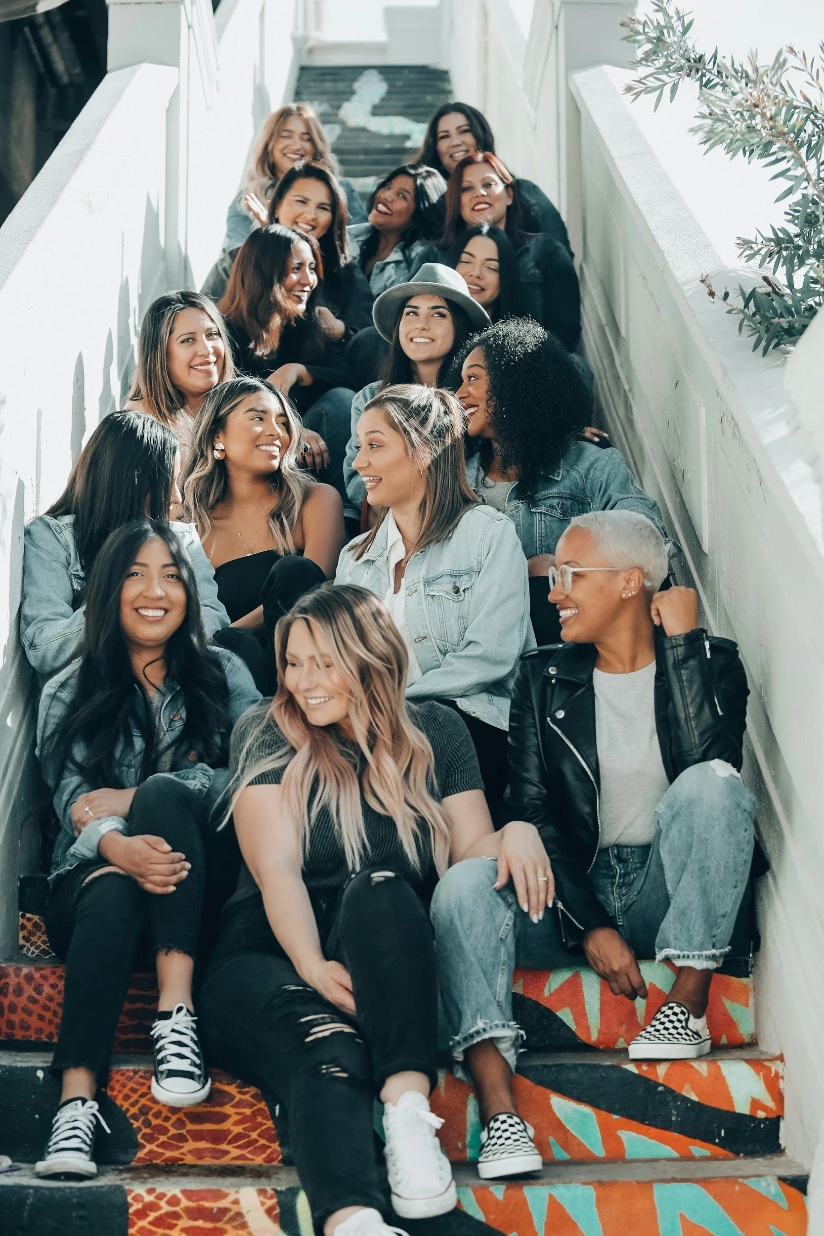 A group of diverse women sitting on stairs indoors, smiling and chatting, some with curly hair, some with straight hair, wearing casual clothing like denim jackets and sneakers, with natural light illuminating the scene.