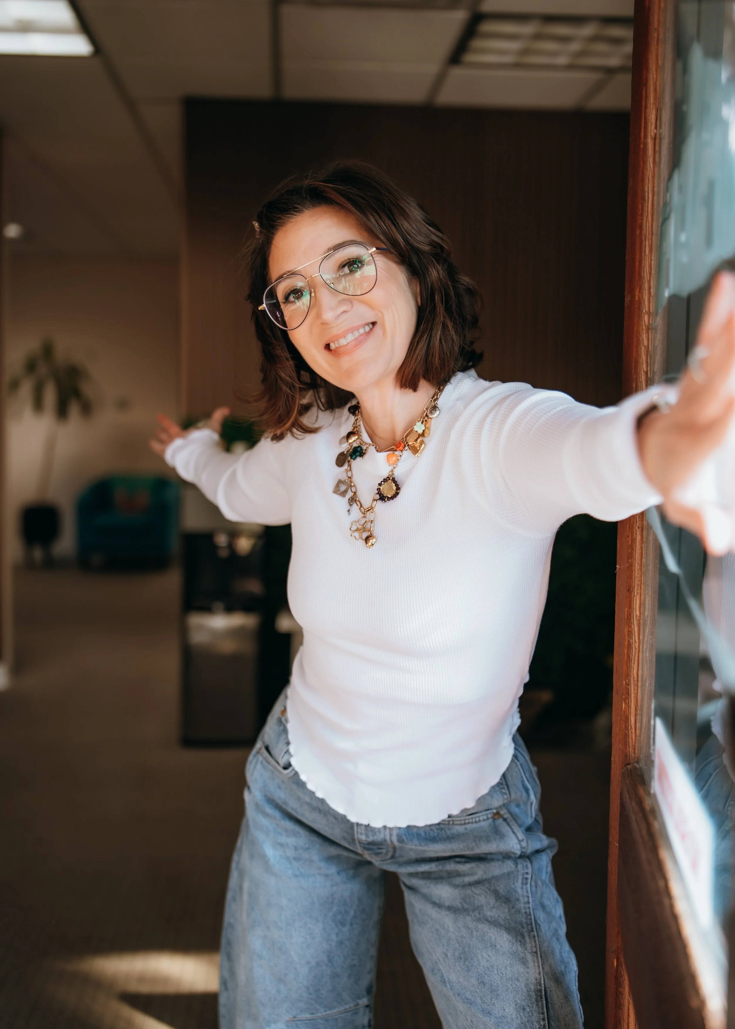 Woman with glasses and jewelry taking a selfie indoors, wearing a white top and blue jeans.