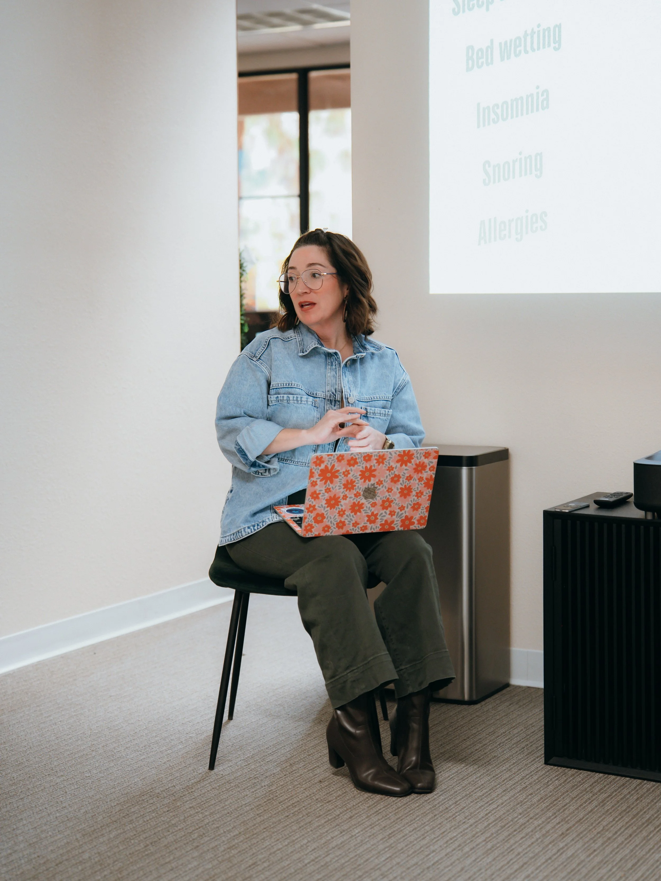 A woman with glasses and shoulder-length dark hair, wearing a denim jacket, dark pants, and brown boots, sitting on a black stool in a room with beige carpet and white walls, next to a small black cabinet and a silver trash bin, with a presentation slide projected on the wall behind her.