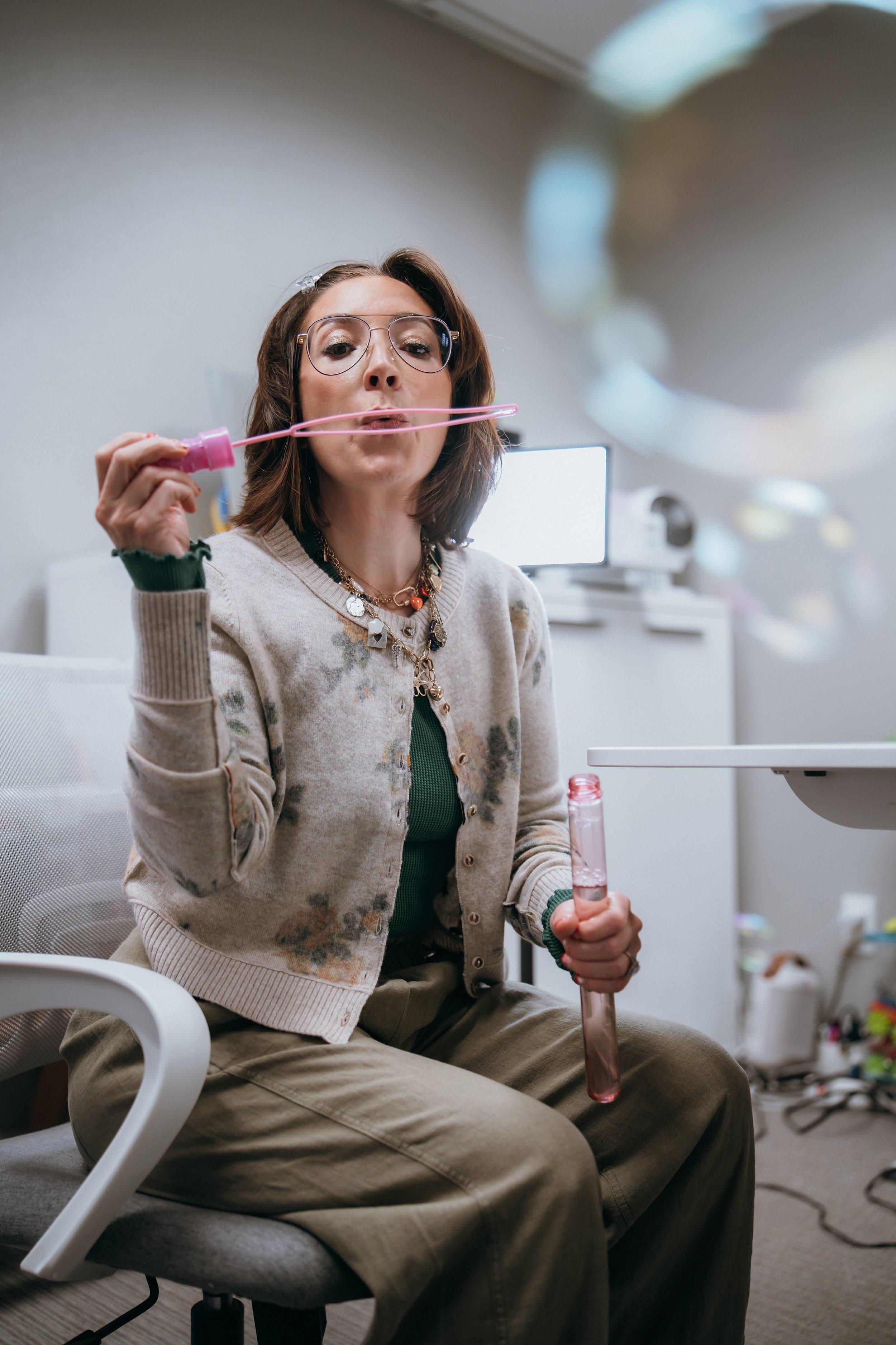 A woman with glasses and layered necklaces is sitting in an office, blowing bubbles with a pink bubble wand. She holds a test tube in her other hand. The background shows a computer monitor and art supplies.