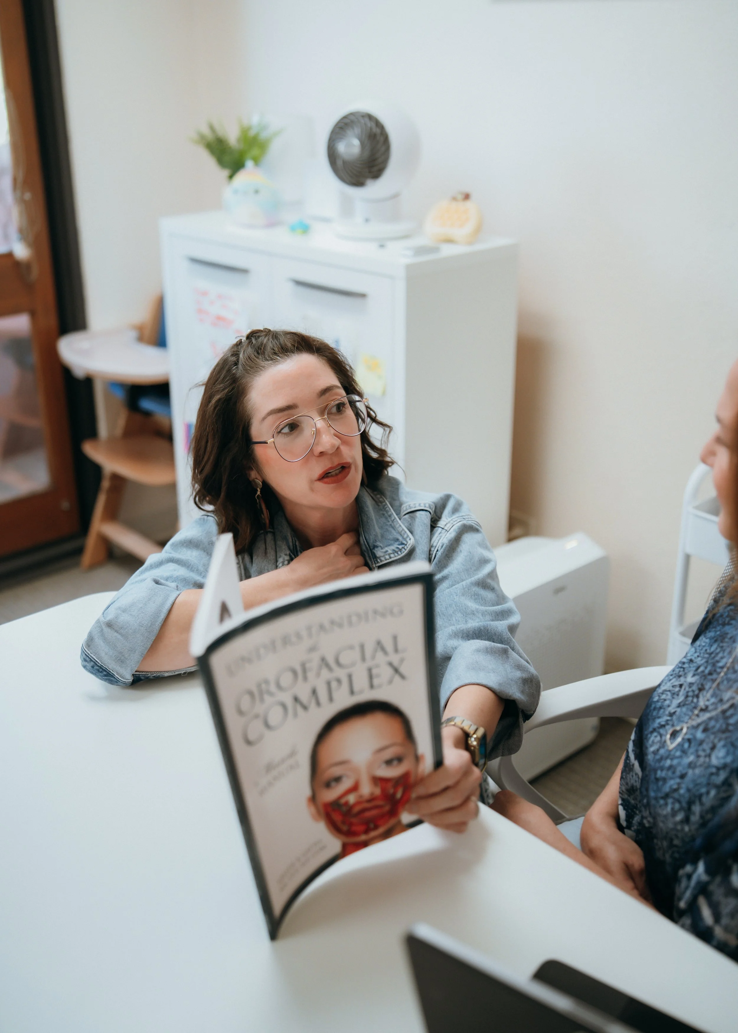 A woman with brown hair and glasses is sitting at a table holding a book about facial recognition technology. She is talking to another person, who is partially visible. In the background, there is a white cabinet with decorations, a small fan, and a potted plant.