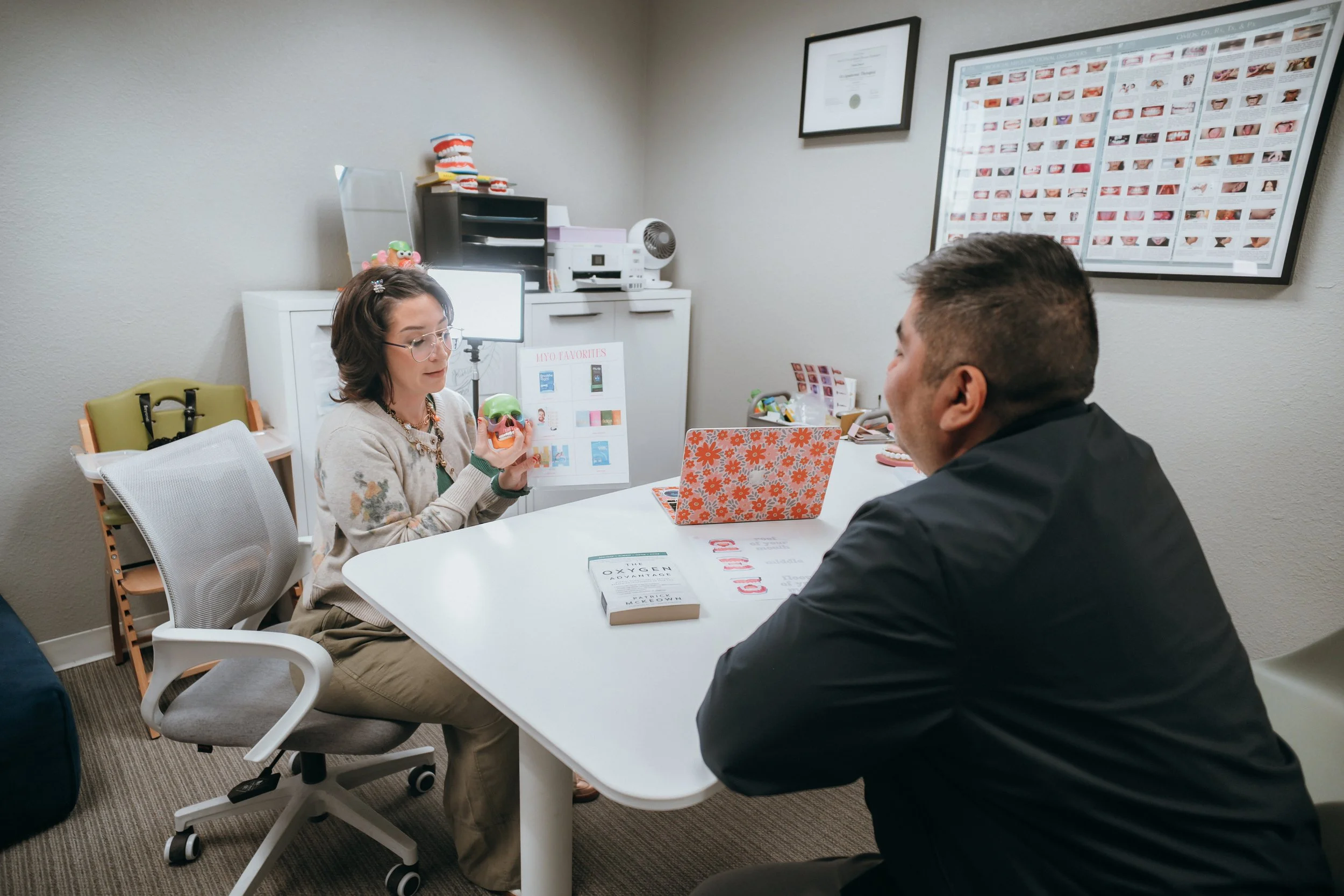 A woman and a man sit across from each other at a white table in a medical office. The woman is holding a model of teeth and discussing dental options. The room has framed certificates, posters, and dental models on the walls and shelves.