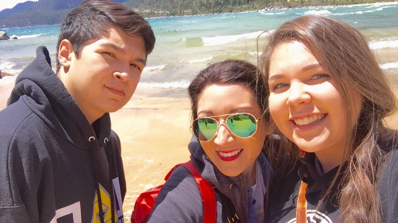 Three people smiling at the camera on a beach with ocean waves and mountains in the background.
