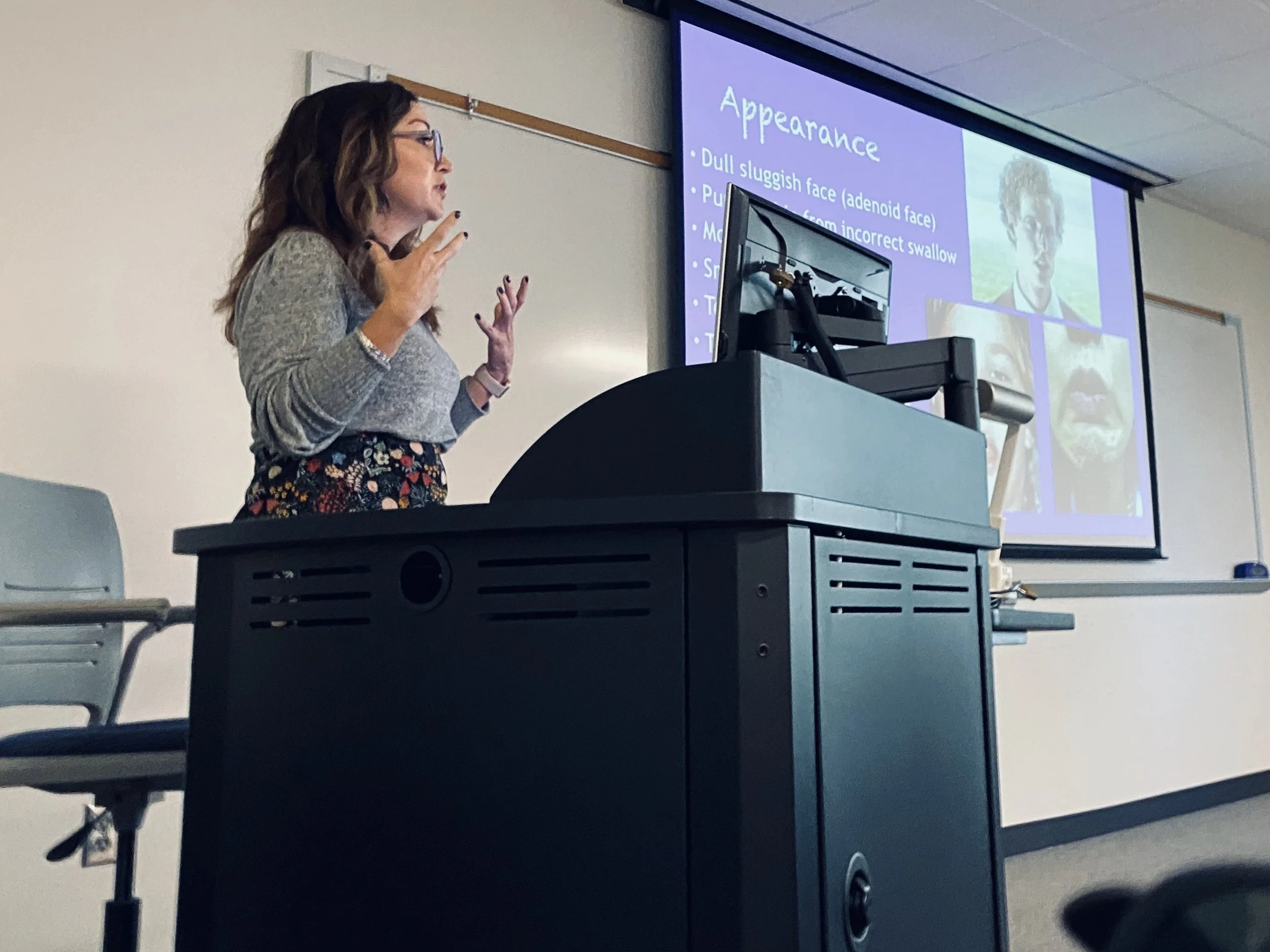 A woman with long dark hair, glasses, and a gray sweater giving a presentation in a classroom. She is standing near a lectern, gesturing with her hands. Behind her, a large screen displays a PowerPoint slide titled 'Appearance' with bullet points about facial features. There is a desktop computer and some cords on the lectern, and chairs are visible in the foreground.
