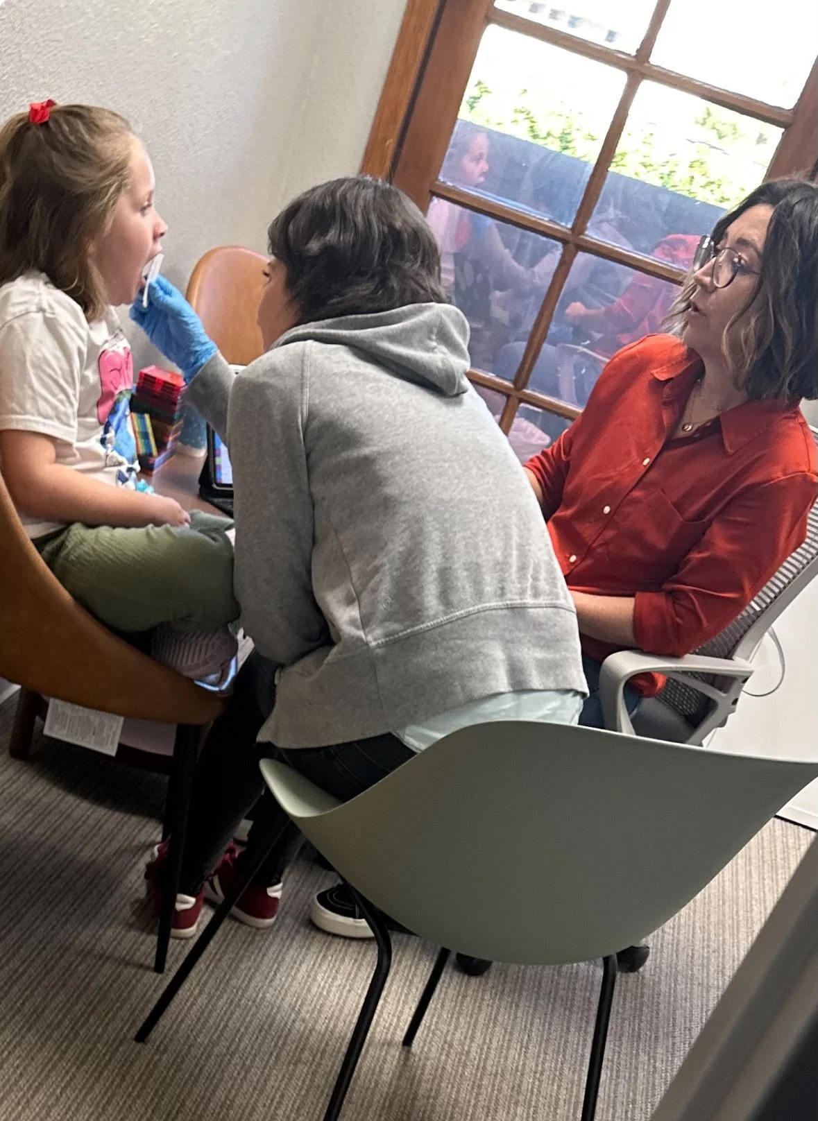 A woman in glasses and a red shirt is sitting at a table with a girl, holding her chin, while another woman in a gray hoodie stands in front of them and takes a Covid-19 test sample.