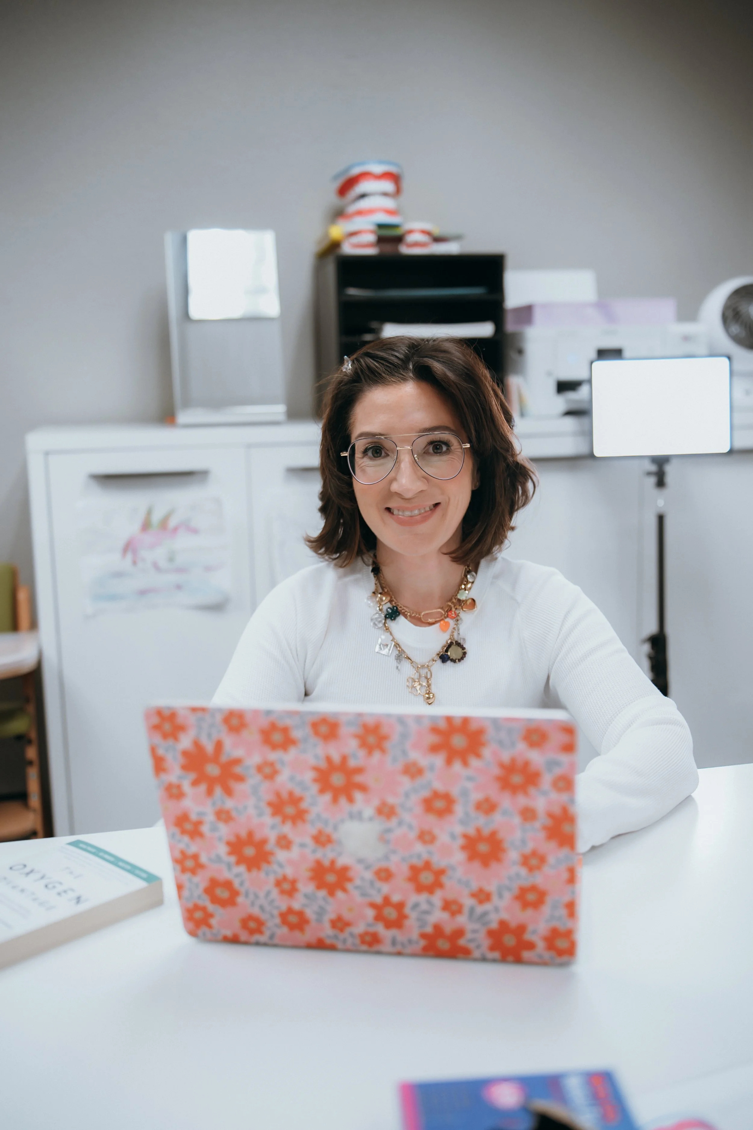 A woman with shoulder-length brown hair, glasses, and a colorful necklace, sitting at a white table, smiling while looking at a colorful floral-patterned laptop. In the background, there is a black shelving unit with a folded towel or blanket on top, a mirror, and some office equipment.