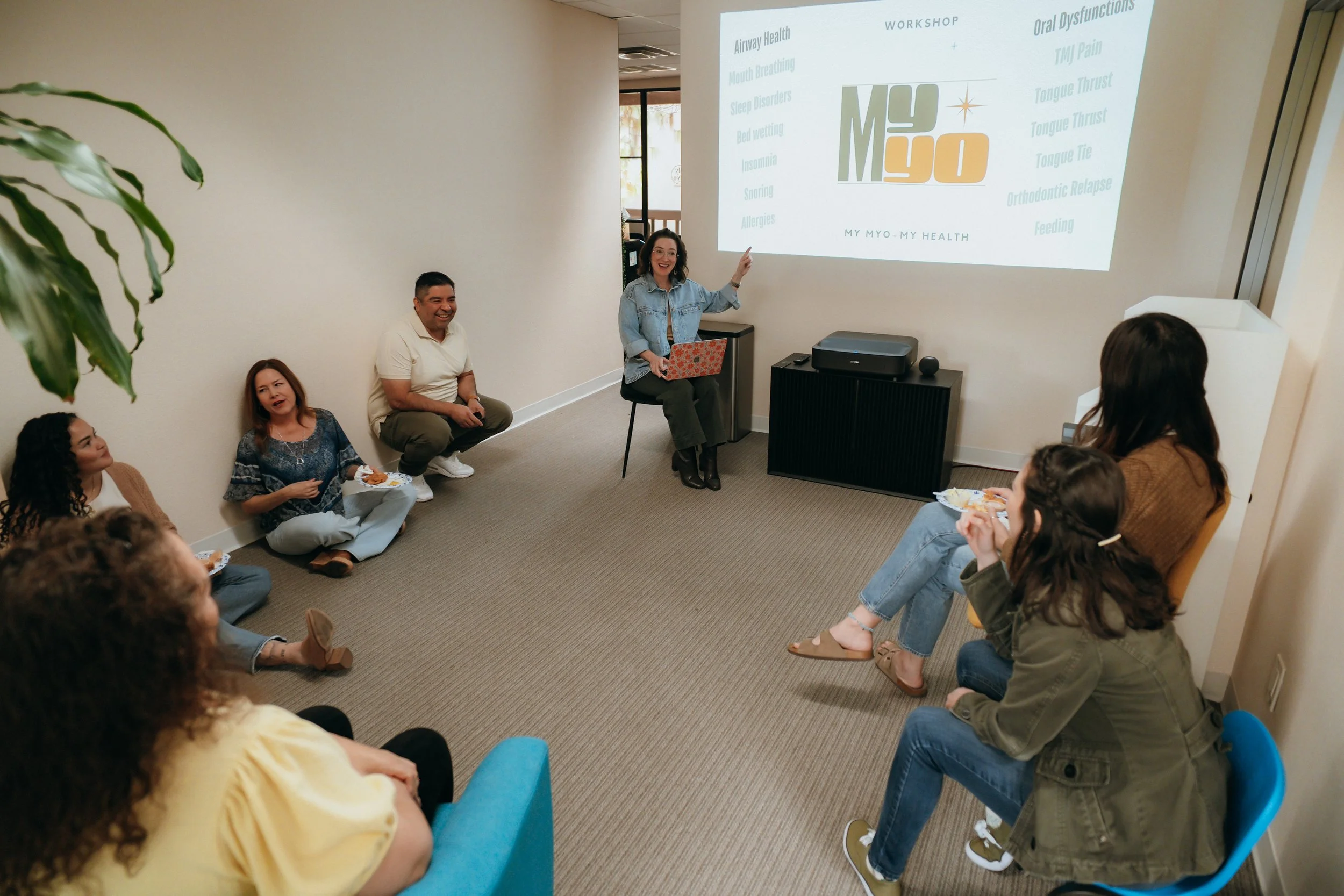 A woman leading a health workshop about MYO MY HEALTH to a group of people seated on the floor and chairs in a meeting room. A projection screen displays workshop topics.
