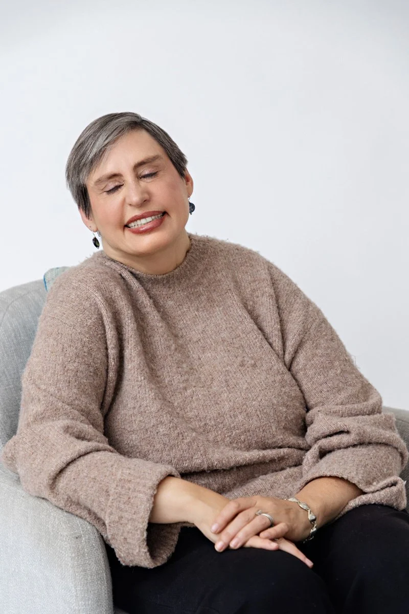 A woman with short gray hair sitting comfortably with her eyes closed and a gentle smile, wearing a light pink sweater and sitting on a light gray chair against a plain white background.