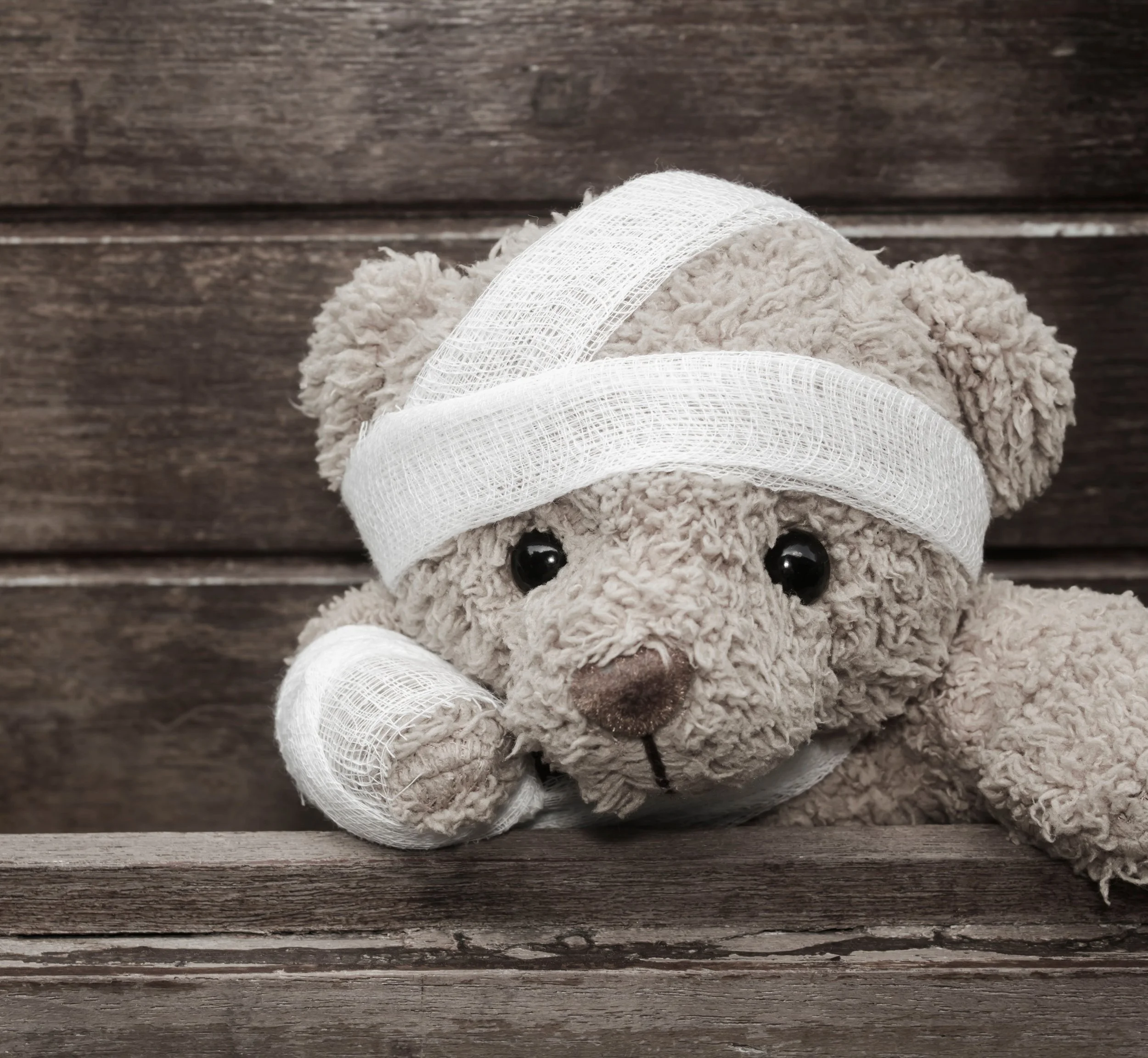 A plush teddy bear with bandages on its head and arm, resting its head on a wooden surface against a wooden background.