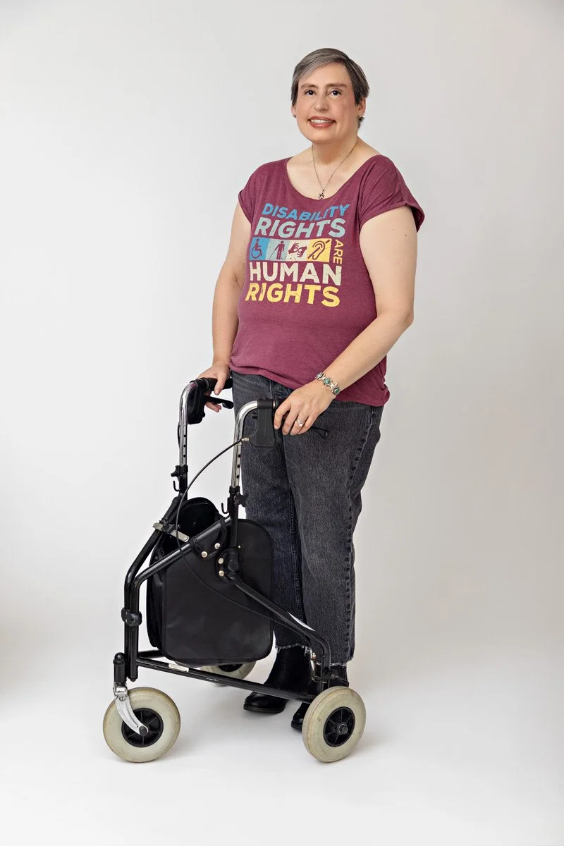 Woman smiling standing with a walker, wearing a maroon T-shirt with Disability Rights are Human Rights written on it, against a plain white background.