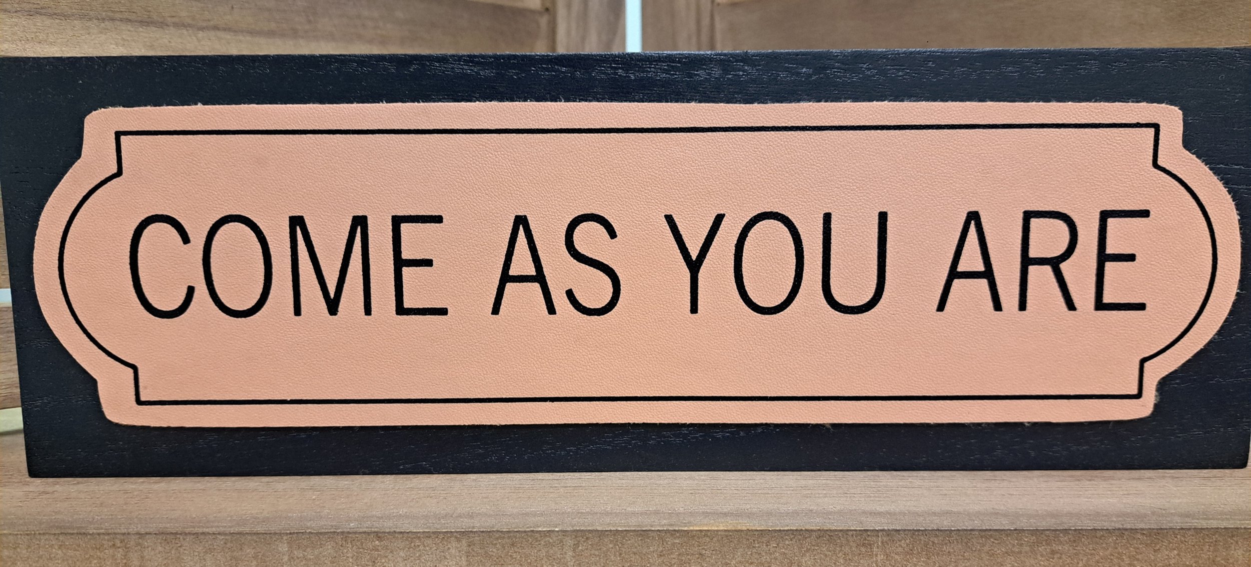 A sign with the words 'Come As You Are' written in black on a pink background, bordered with a black outline.