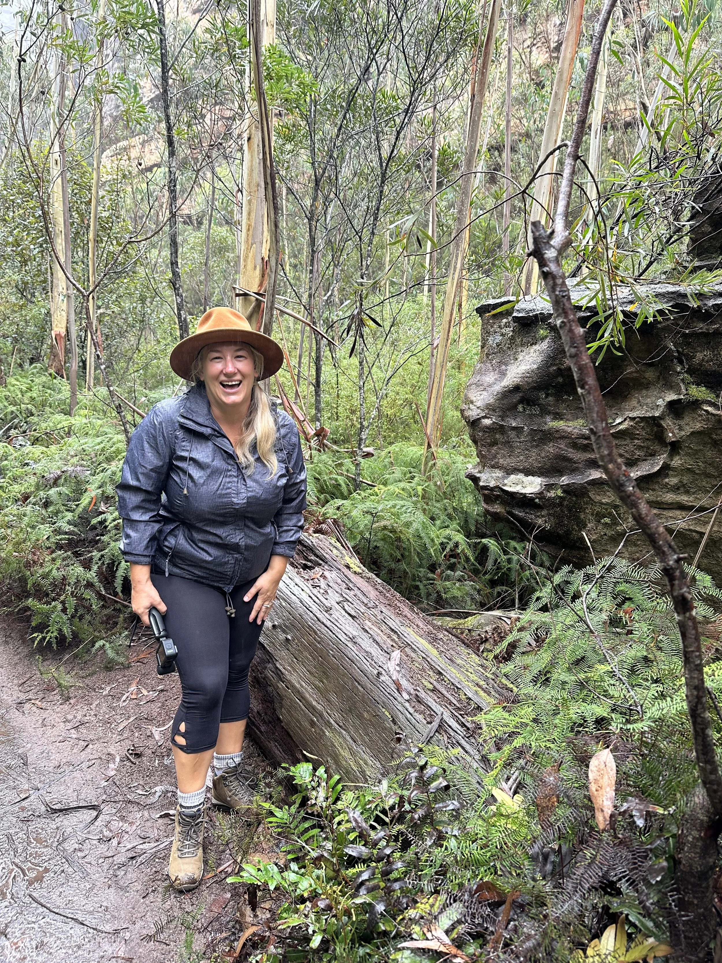 A woman in outdoor gear, including a brown hat, black jacket, and black leggings, standing on a dirt trail in a lush forest with green ferns and trees, smiling and holding a small object in her right hand.