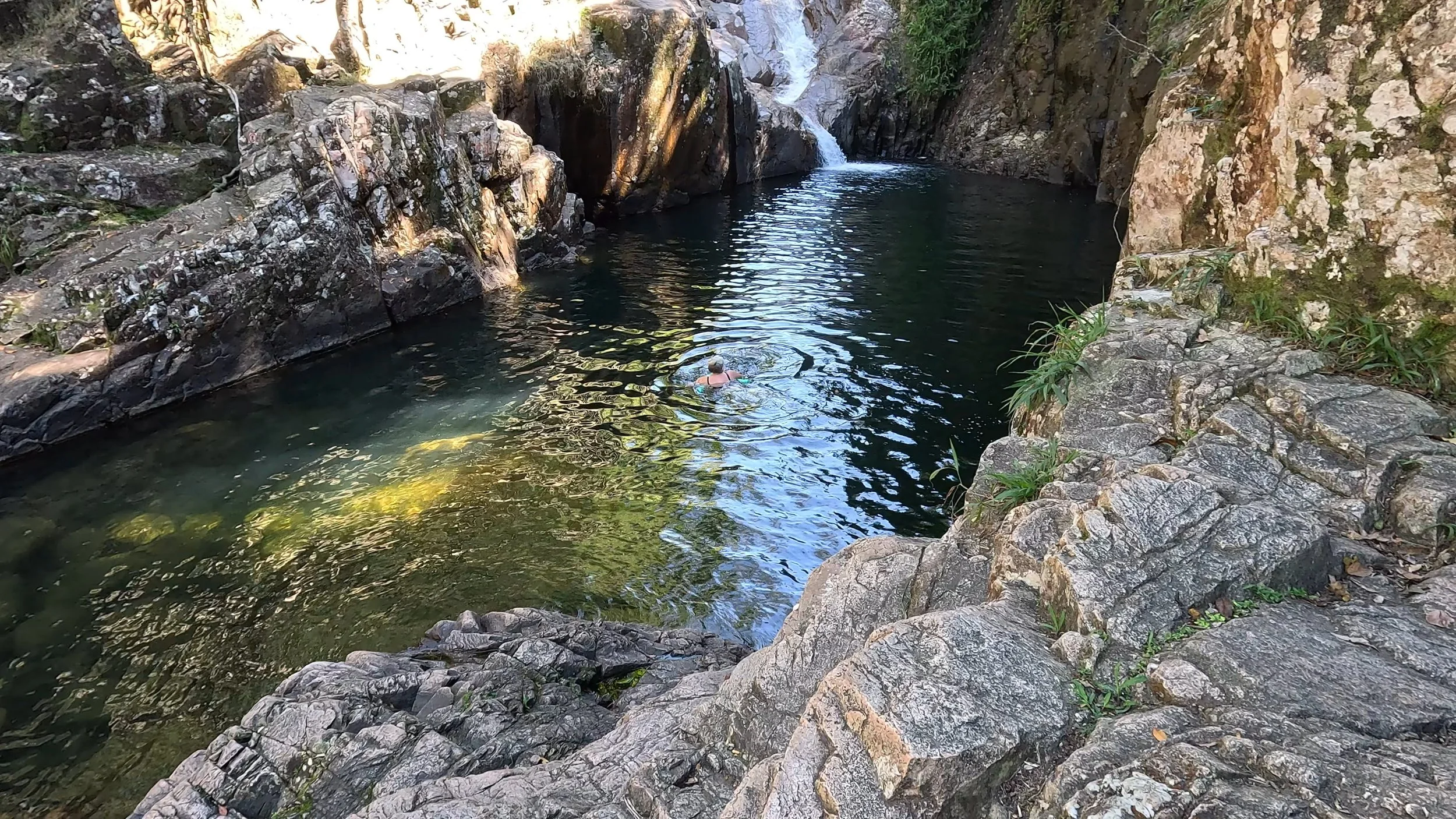 A person swimming in a natural rocky water pool with a small waterfall in the background, surrounded by large rocks and greenery.