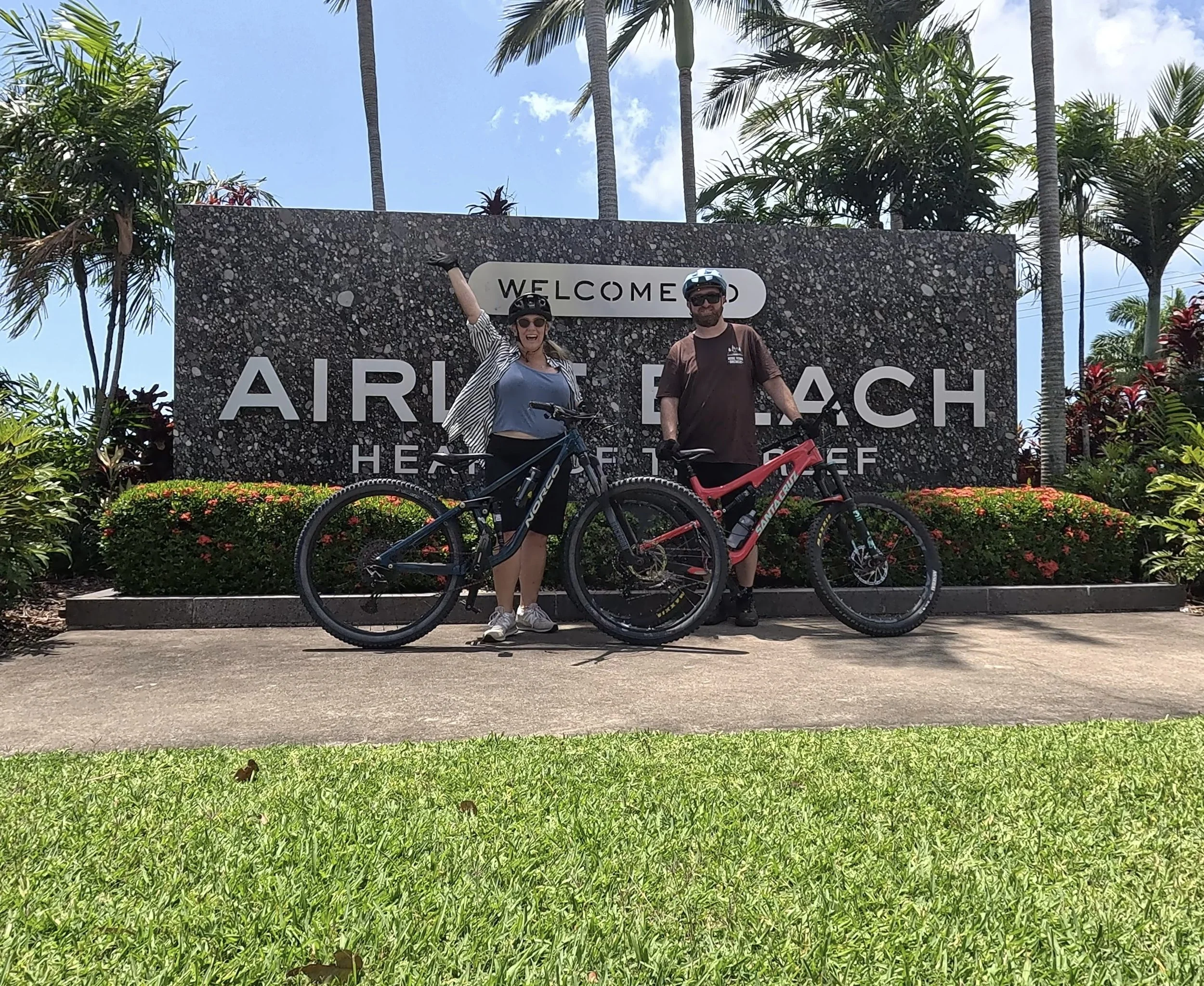 Husband and Wife in cycling gear standing with their bicycles in front of a large sign that reads "Welcome to Airlie Beach." The background includes palm trees, flowering plants, and a blue sky.