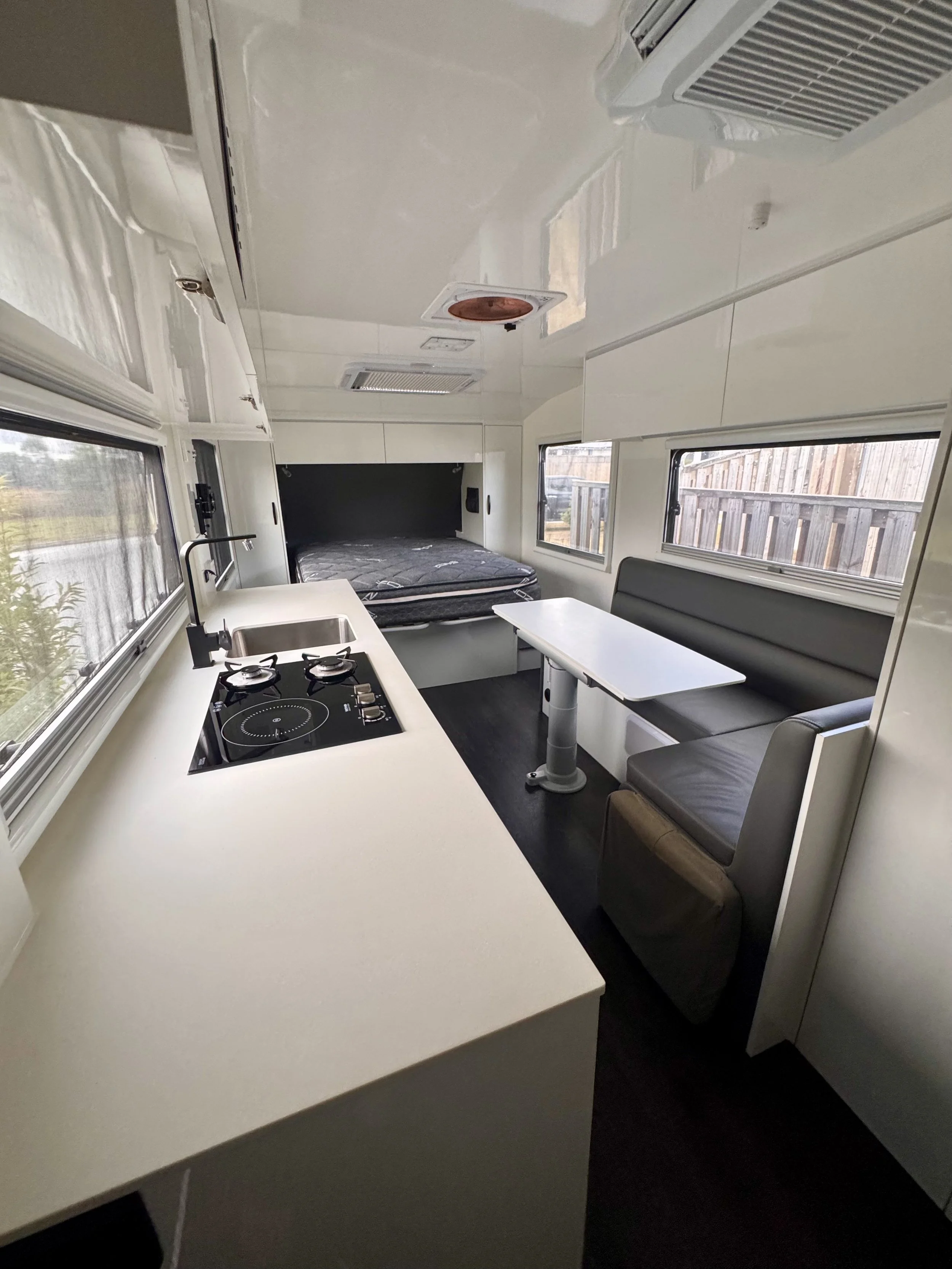 Interior of a compact living space, featuring a kitchen with a white countertop and a two-burner stove, a sleeping area with a mattress, and a small dining area with a bench and a table, all illuminated by natural light from three windows, with a vie