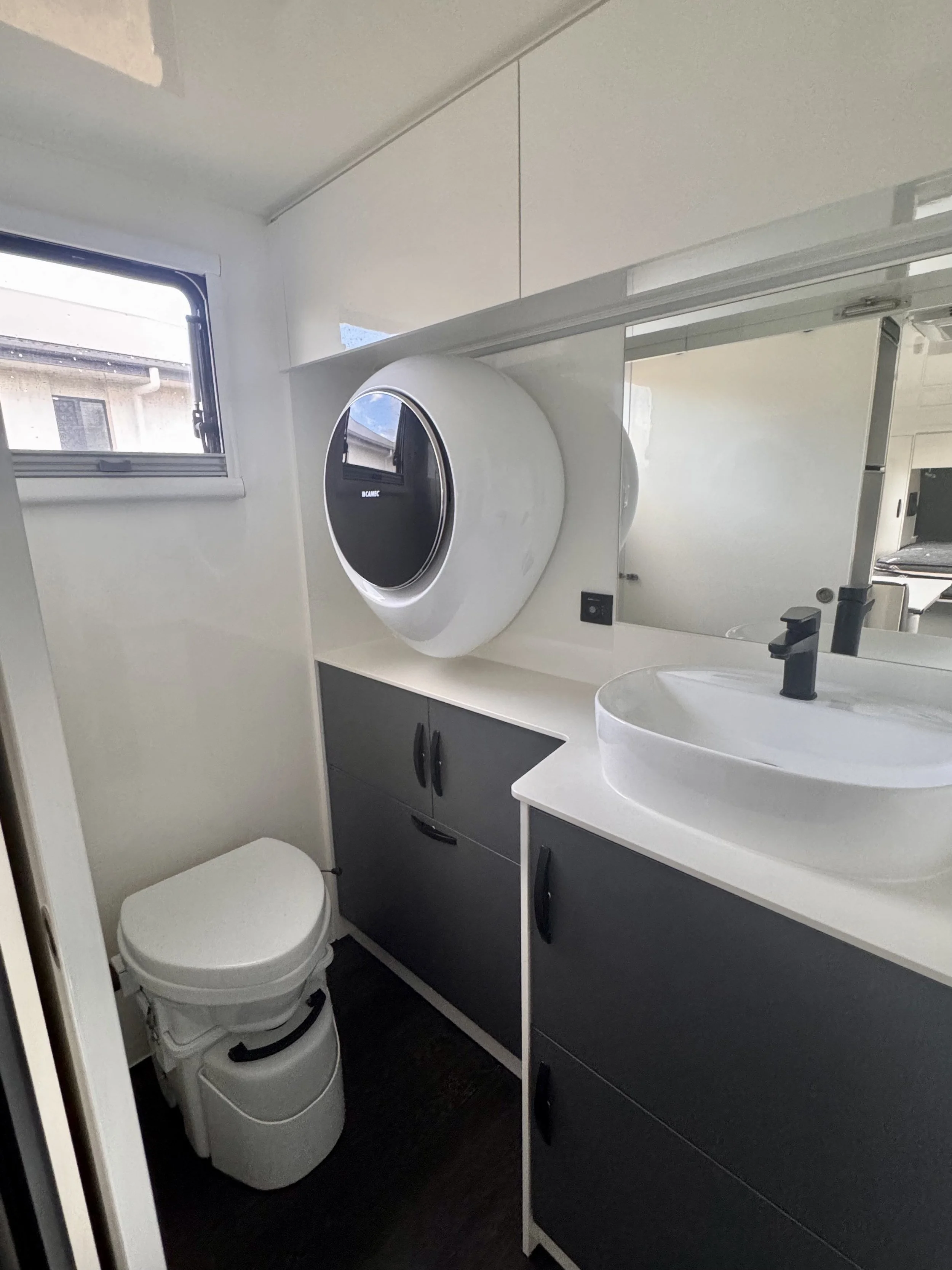 Compact bathroom with a small window, a white toilet, a round vessel sink, a black faucet, a mirror, and dark gray cabinets.