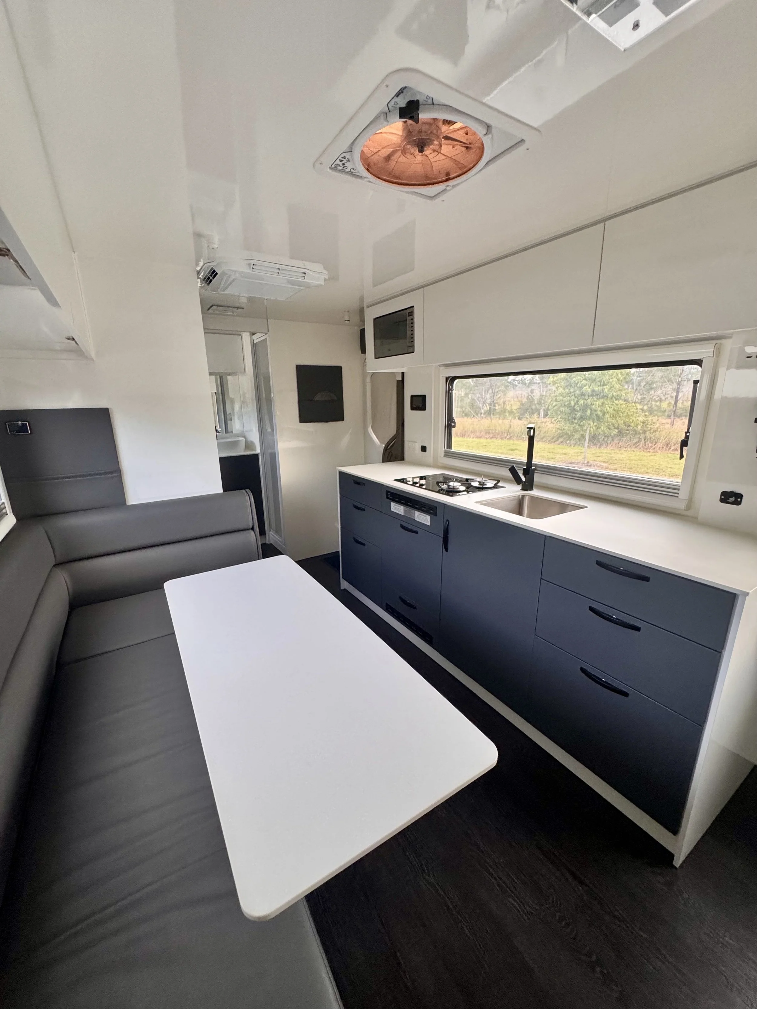 Interior of a caravan living space with a white table, a gray cushioned seating area, and a modern kitchen with grey cabinets, a small sink, stove, and window showing a green outdoor landscape.