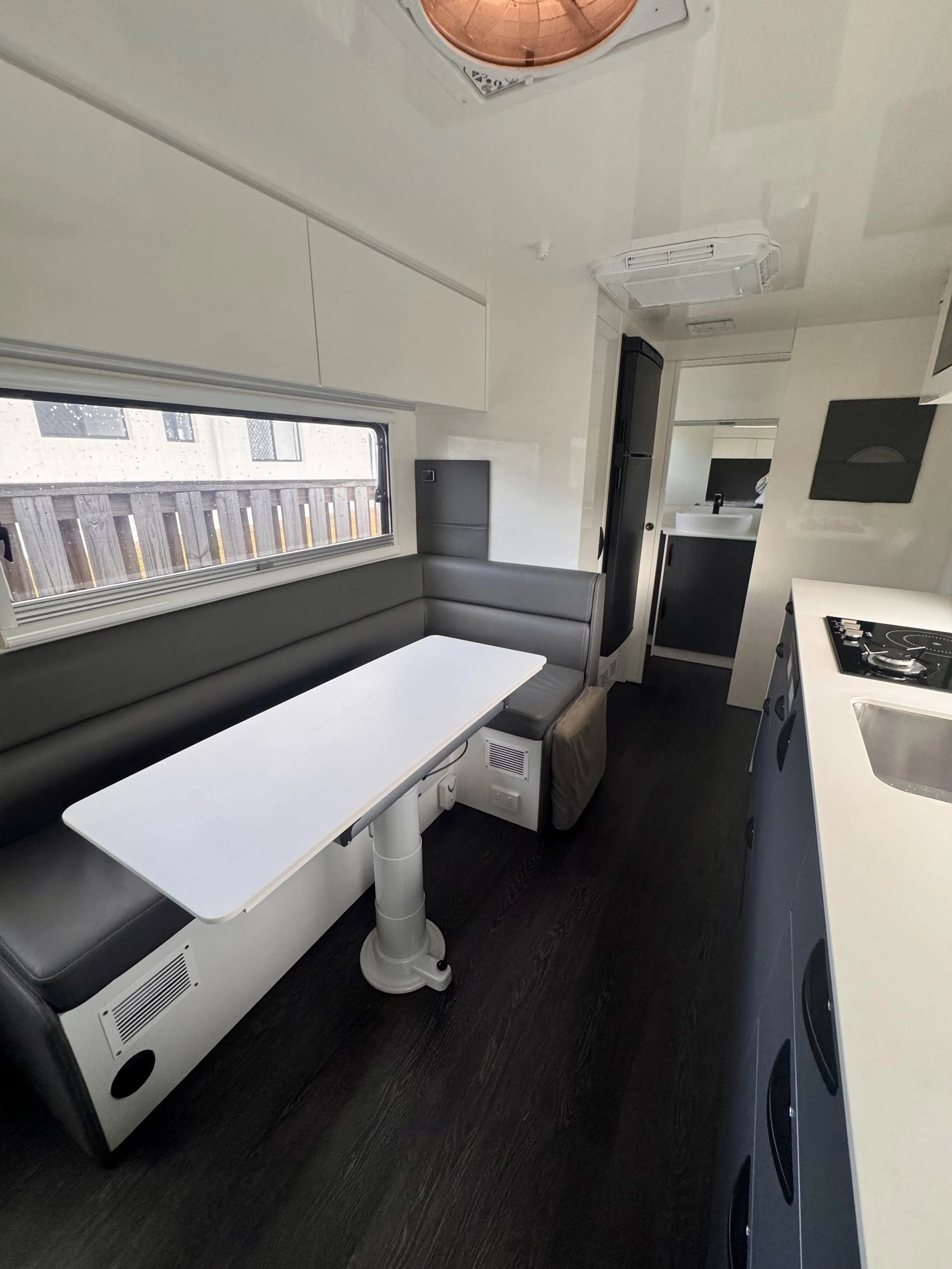 Interior of a compact mobile home or RV kitchen and dining area, featuring a white table with a seating bench, a small kitchenette with a black refrigerator, a sink, a stovetop, a window with blinds, and ceiling air conditioning units.