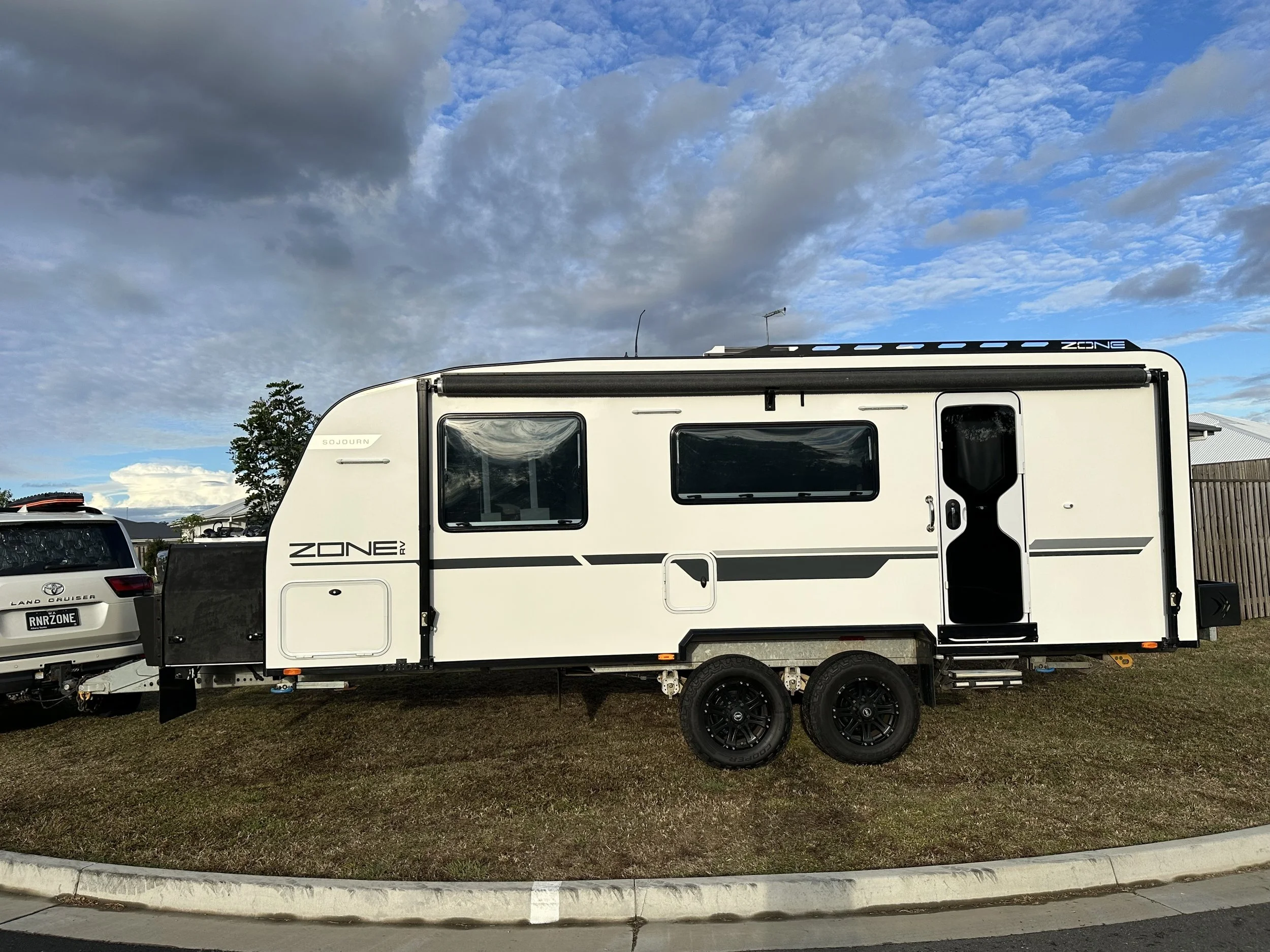 A white caravan with black accents parked on grass near a curb, with a cloudy sky and a tree in the background.