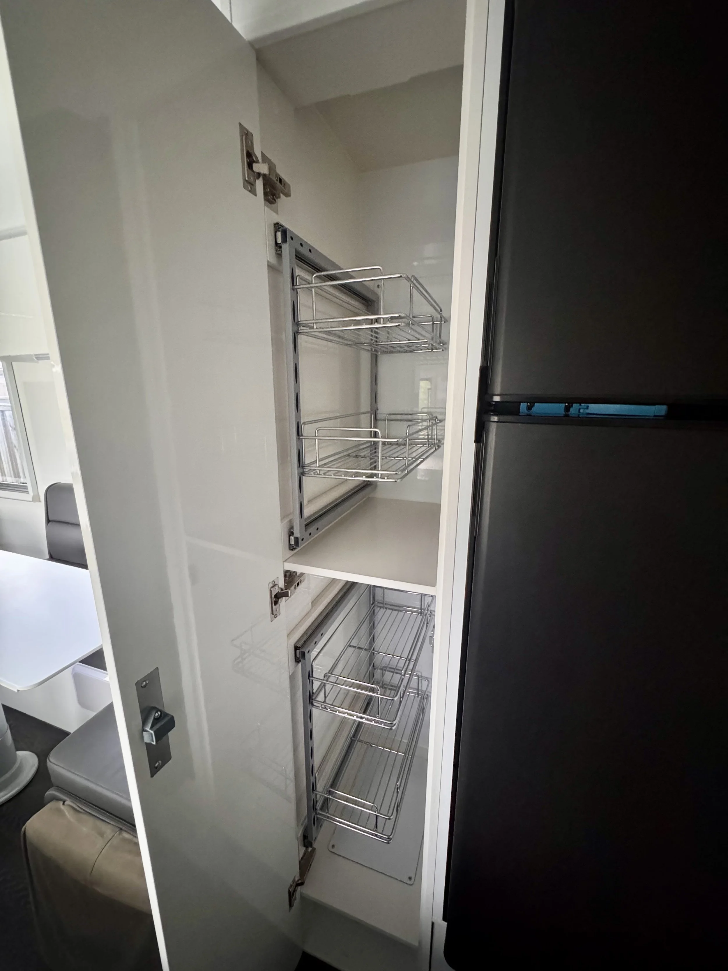 Open kitchen pantry cabinet with pull-out wire baskets, next to a black refrigerator.