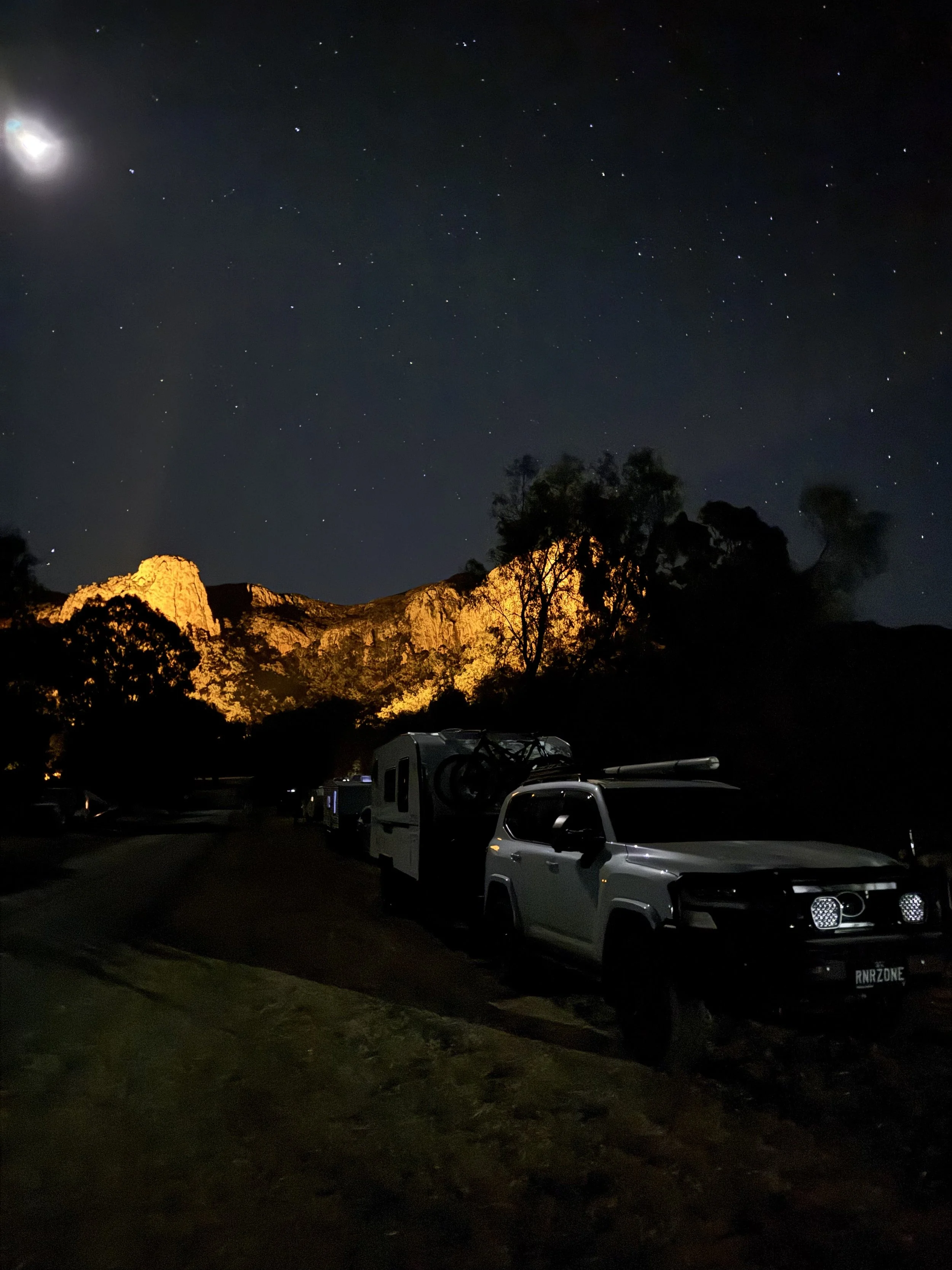 Nighttime scene with a starry sky, illuminated mountains in the background, and parked vehicles including an SUV and a caravan in the foreground.