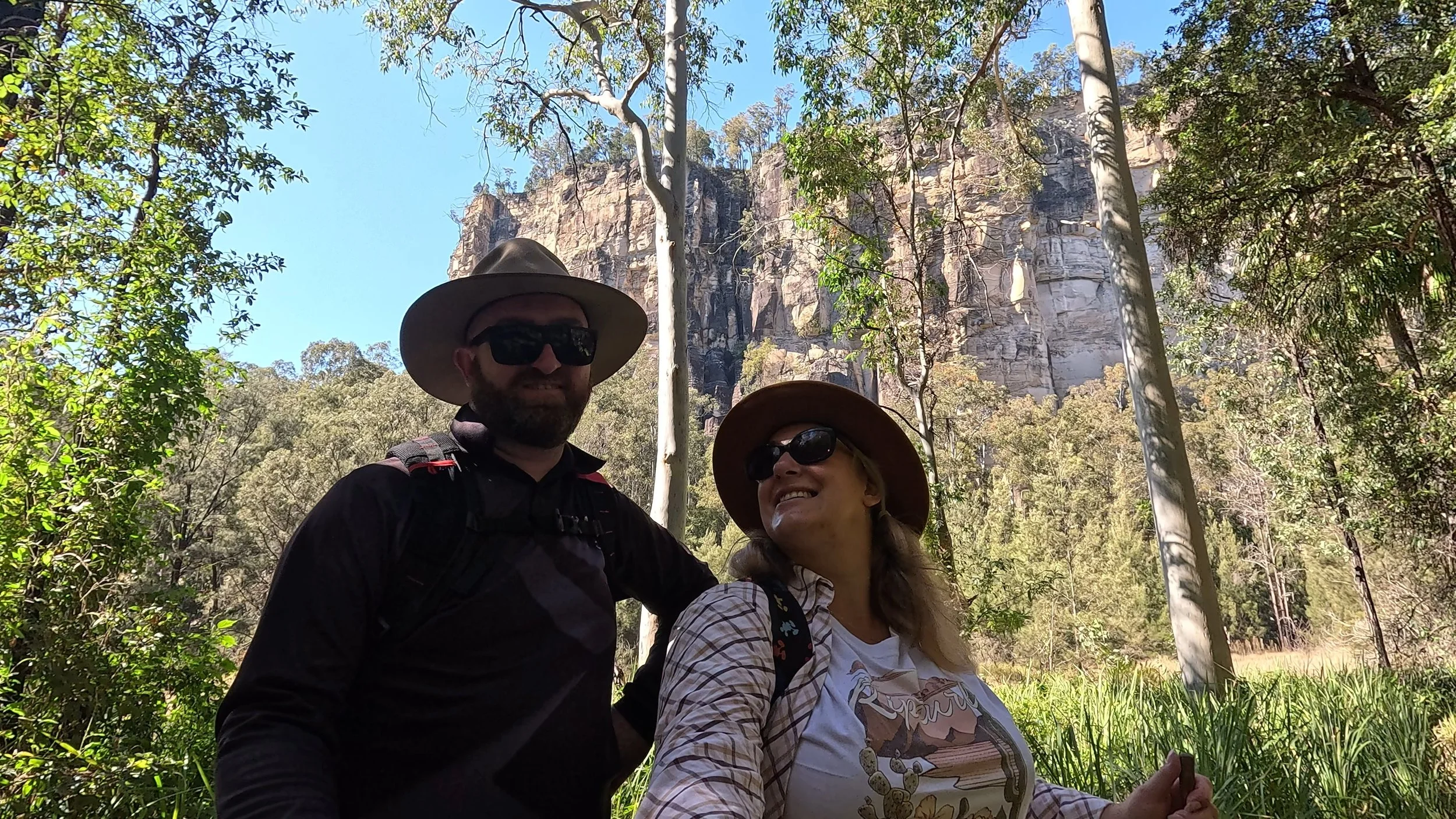 A man and woman hiking in a forest, wearing hats and sunglasses, smiling. The background features tall trees and a large rocky cliff under a clear blue sky.